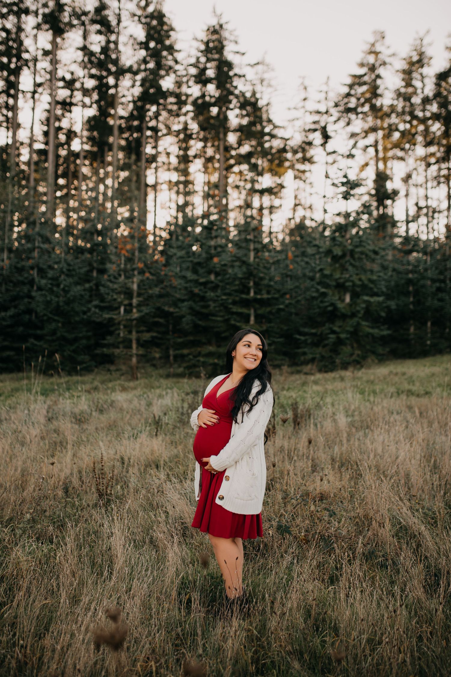 Smiling pregnant woman in a red dress holding her belly in a forest meadow.