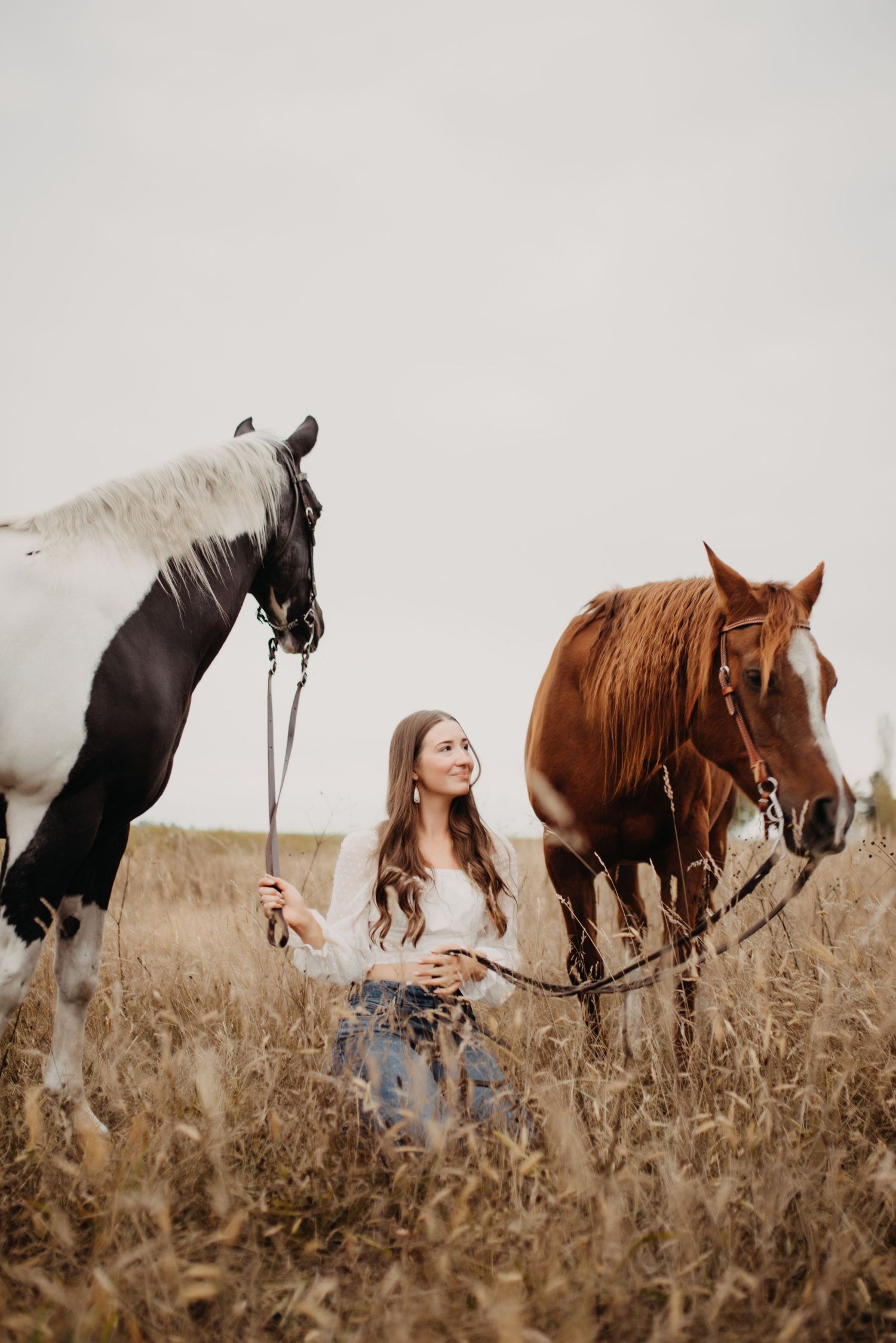 Young woman kneels between two horses in a golden wheat field on an overcast day.