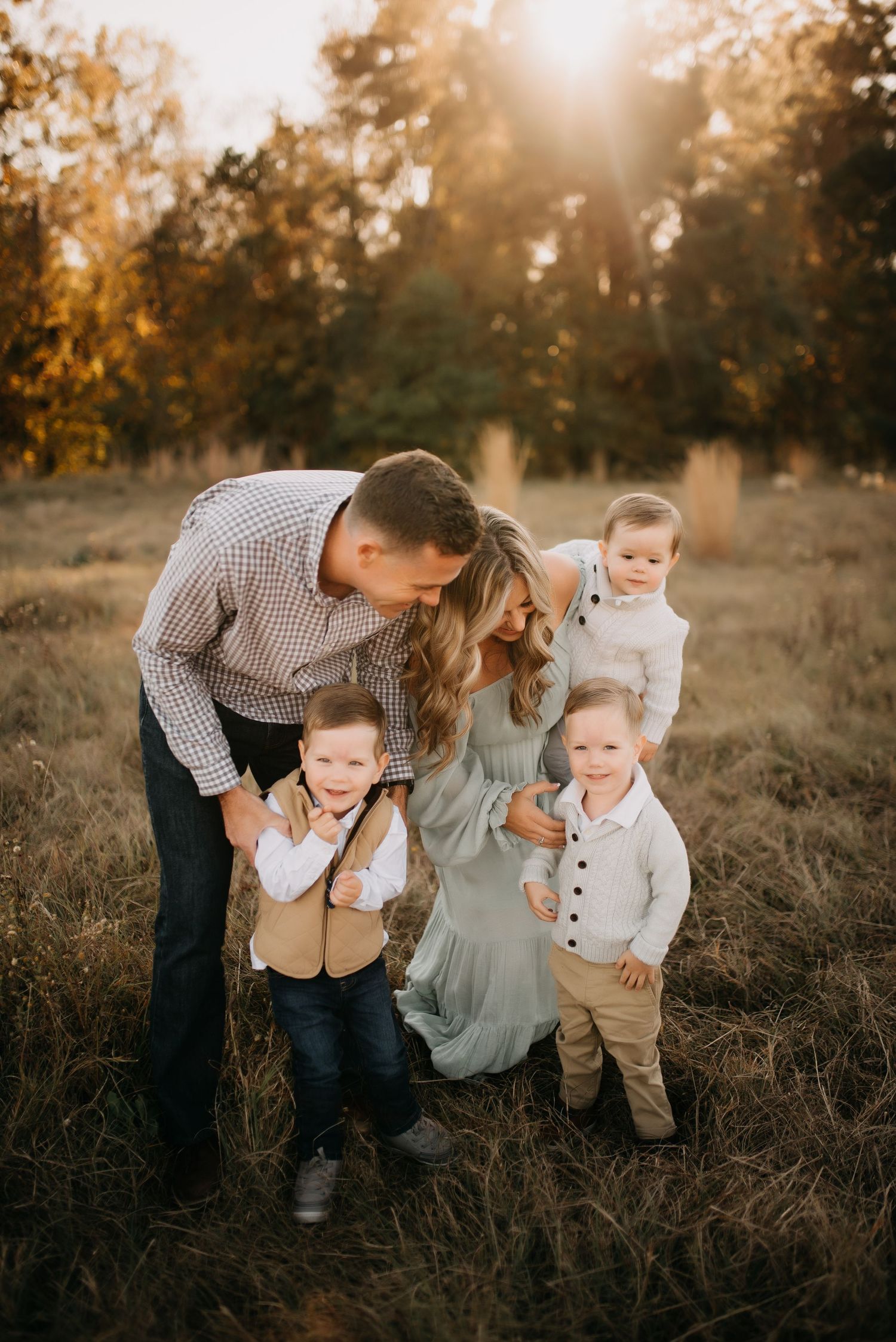 Family of five with three toddler boys posing in golden sunlit field at sunset.