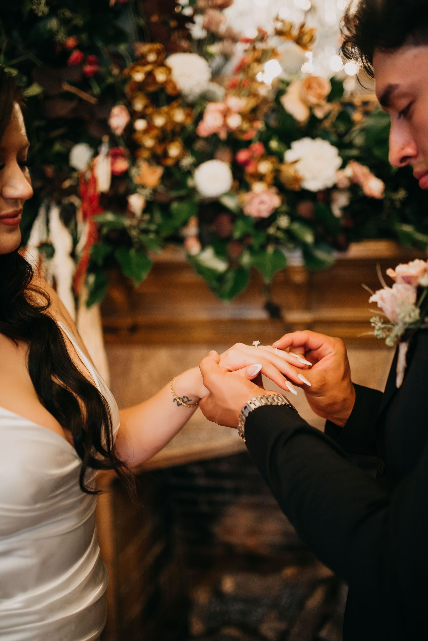 Groom places wedding ring on brides finger at romantic ceremony with lush floral backdrop.