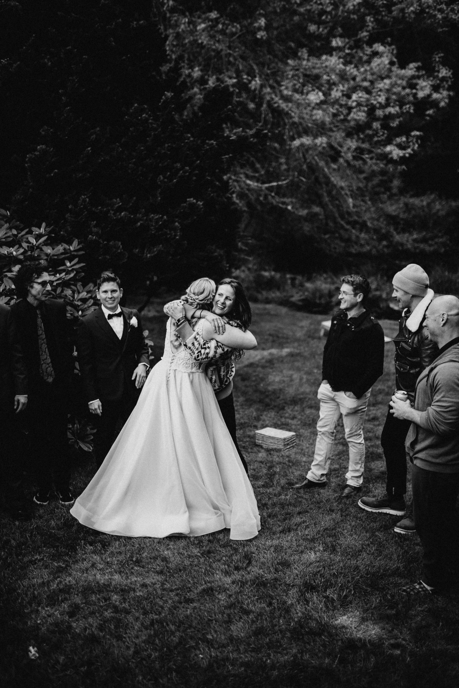 Black and white photo of bride hugging a guest while wedding party looks on in a garden setting.