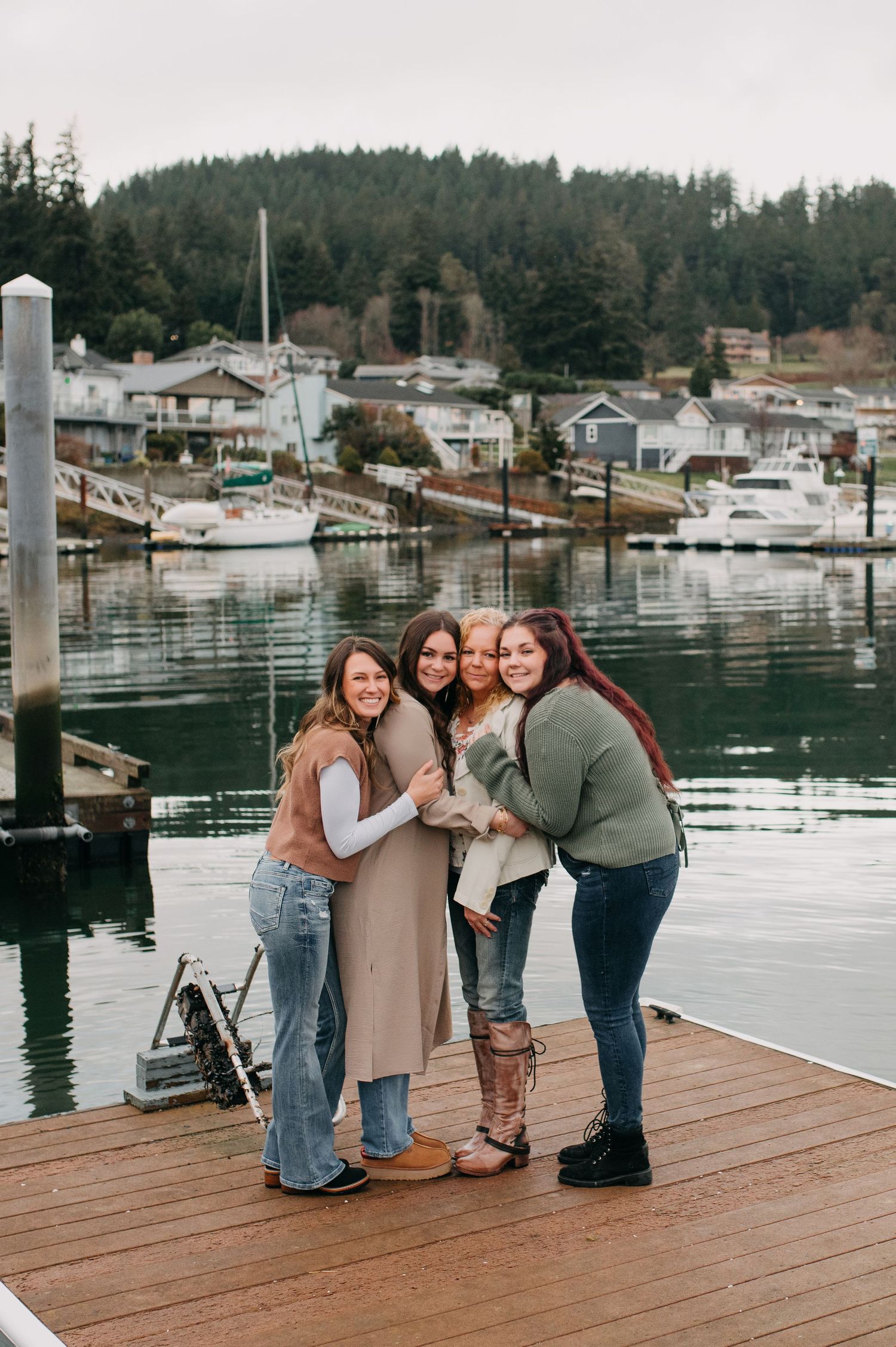 Four women hugging and smiling on a wooden dock with a marina and forested hillside behind them.
