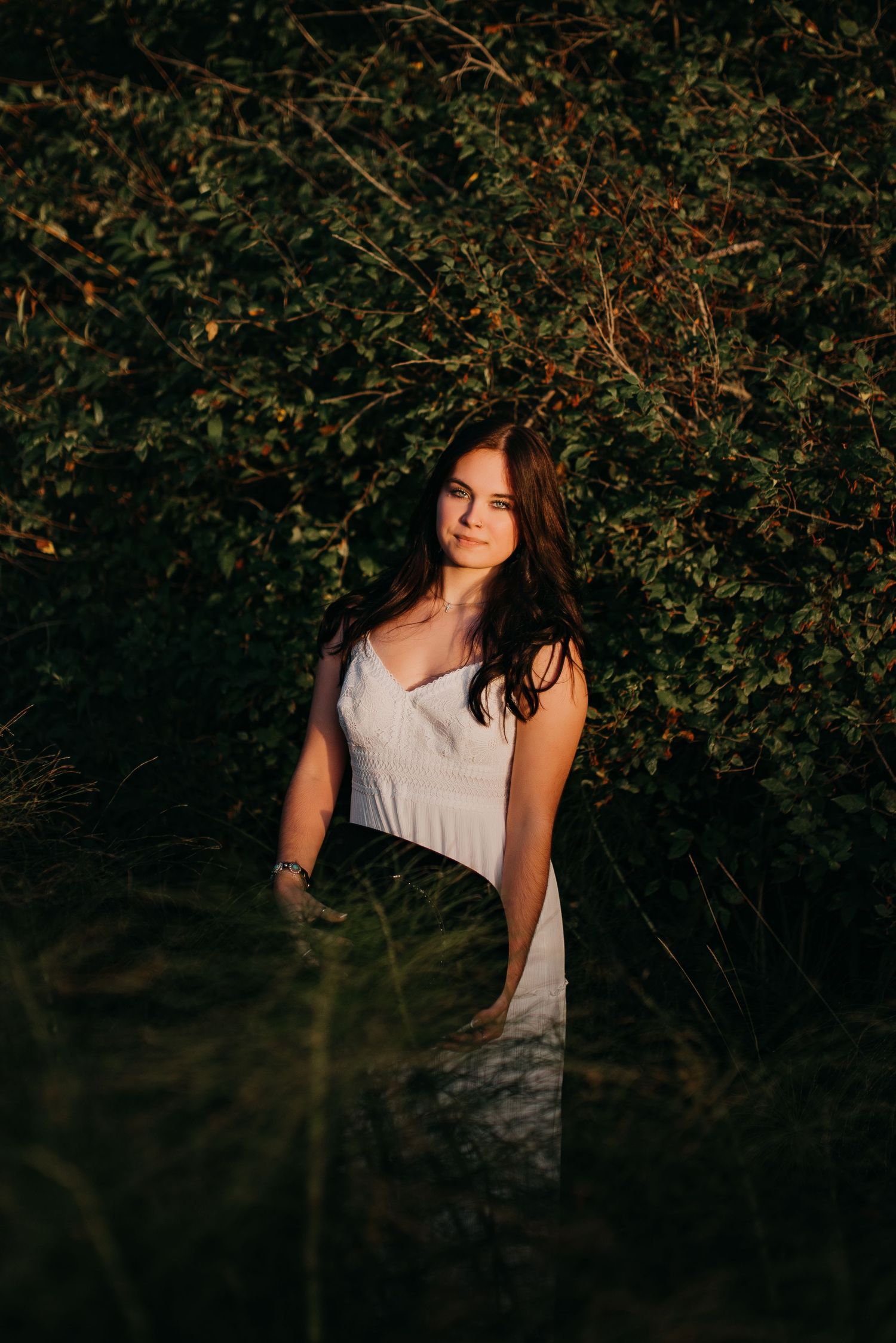 Young woman in white dress standing among lush green bushes in golden hour sunlight.