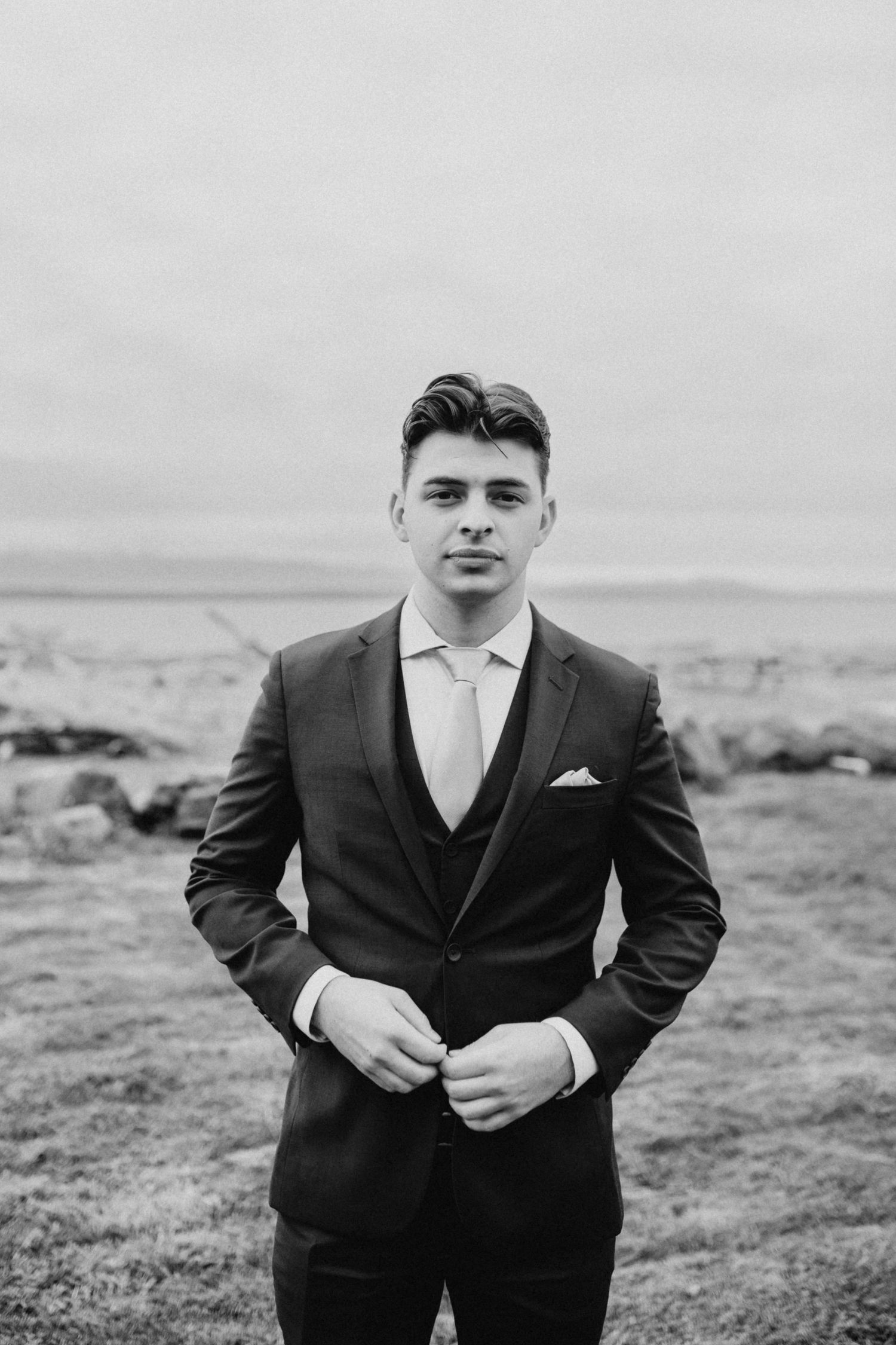 Black and white portrait of a well-dressed groom in a three-piece suit outdoors by the water.