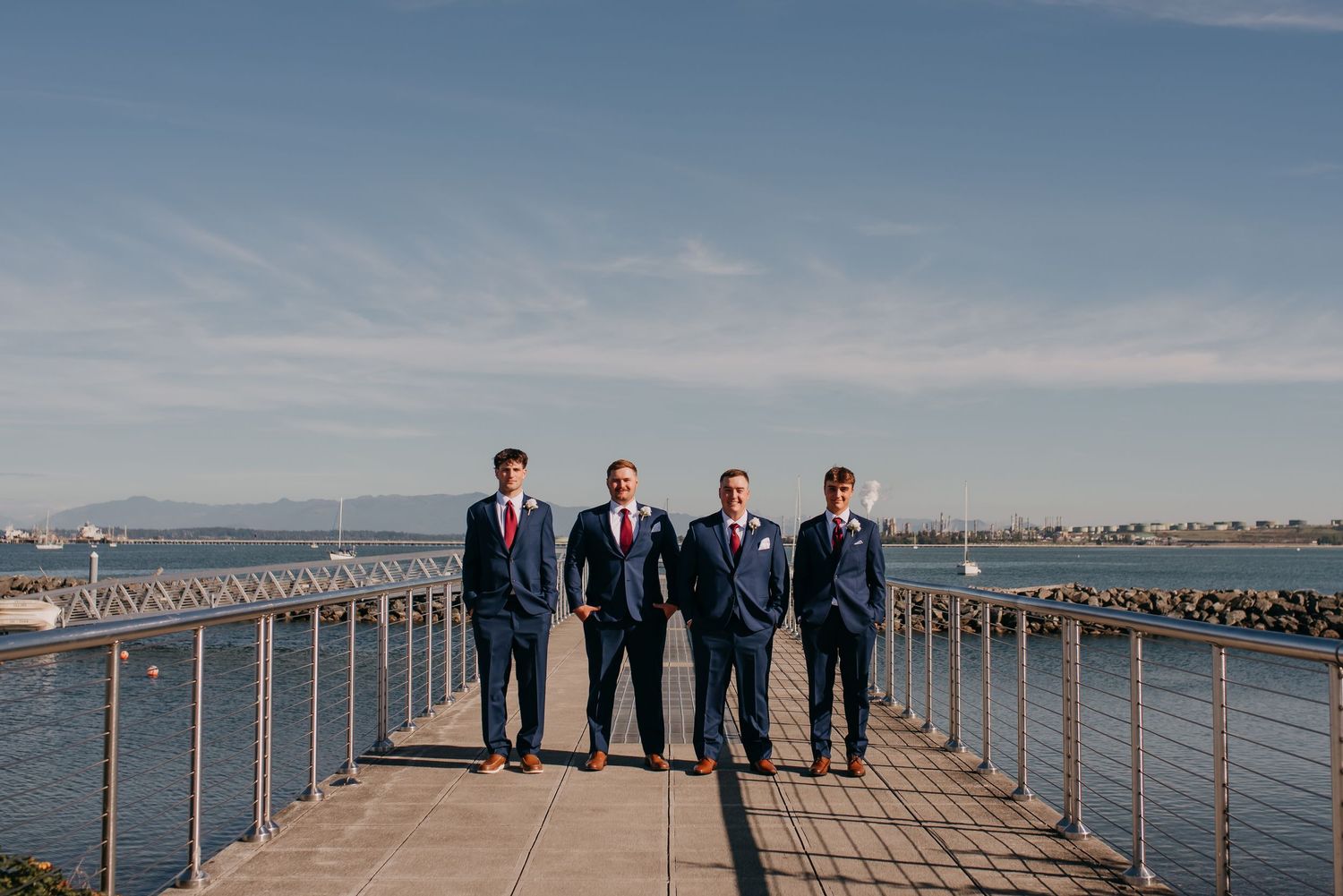 Four groomsmen in navy suits walk confidently along a waterfront pier under a bright blue sky.
