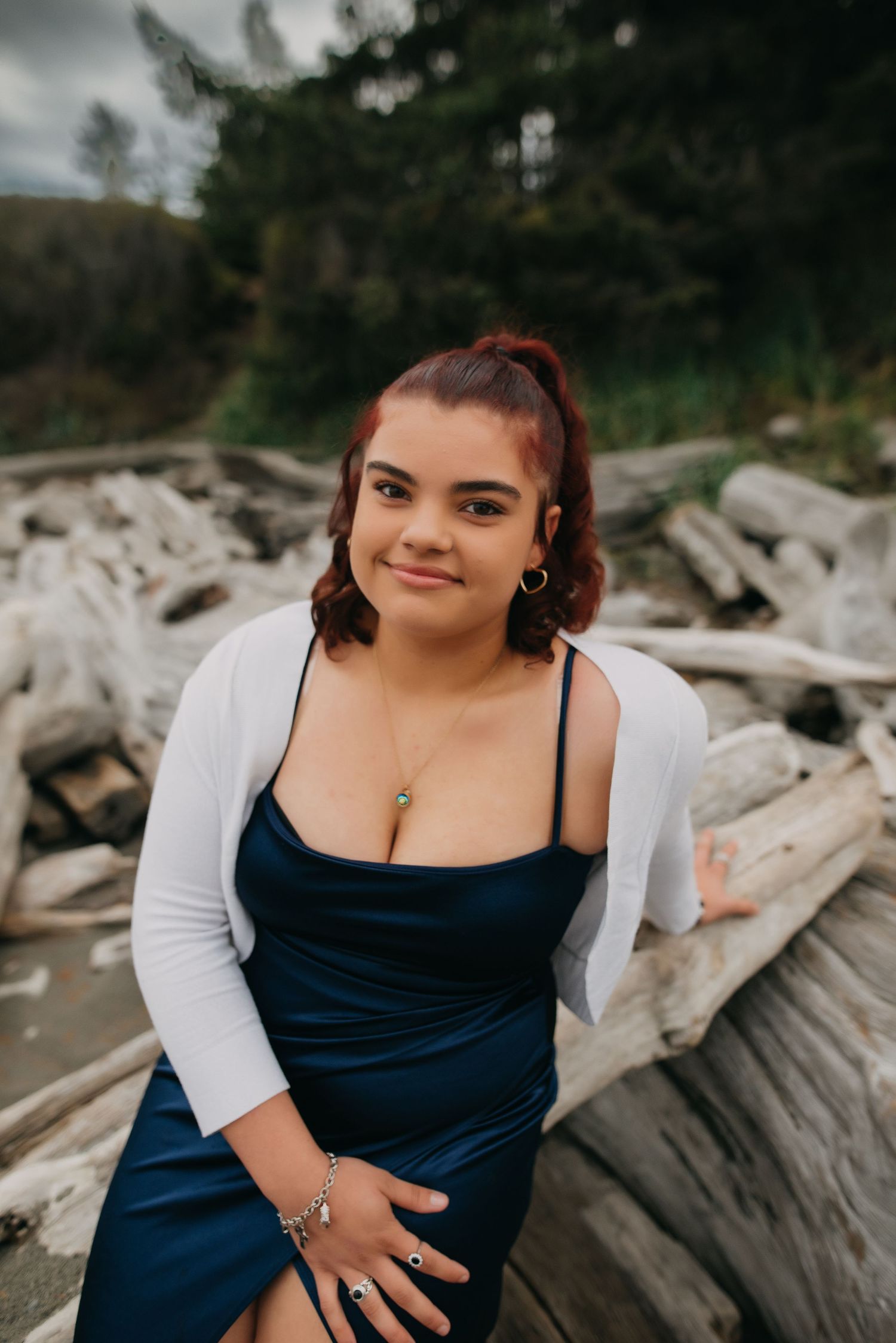 Senior photoshoot with a young woman in blue satin dress and white cardigan sitting on driftwood at a Rosario Park beach.