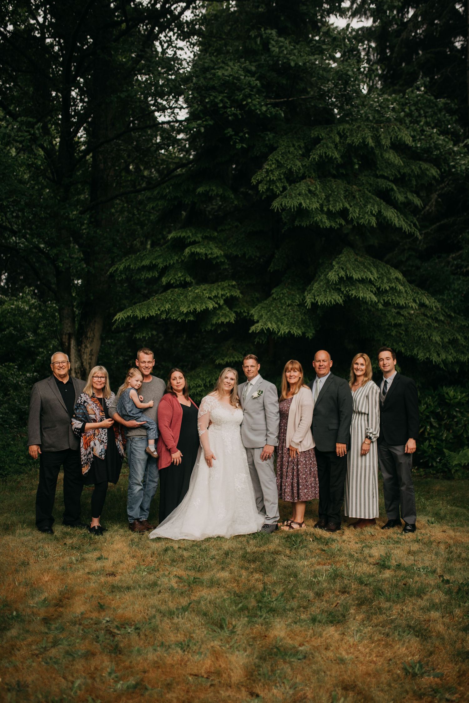 Wedding couple poses with family group in front of towering evergreen trees on a green lawn.