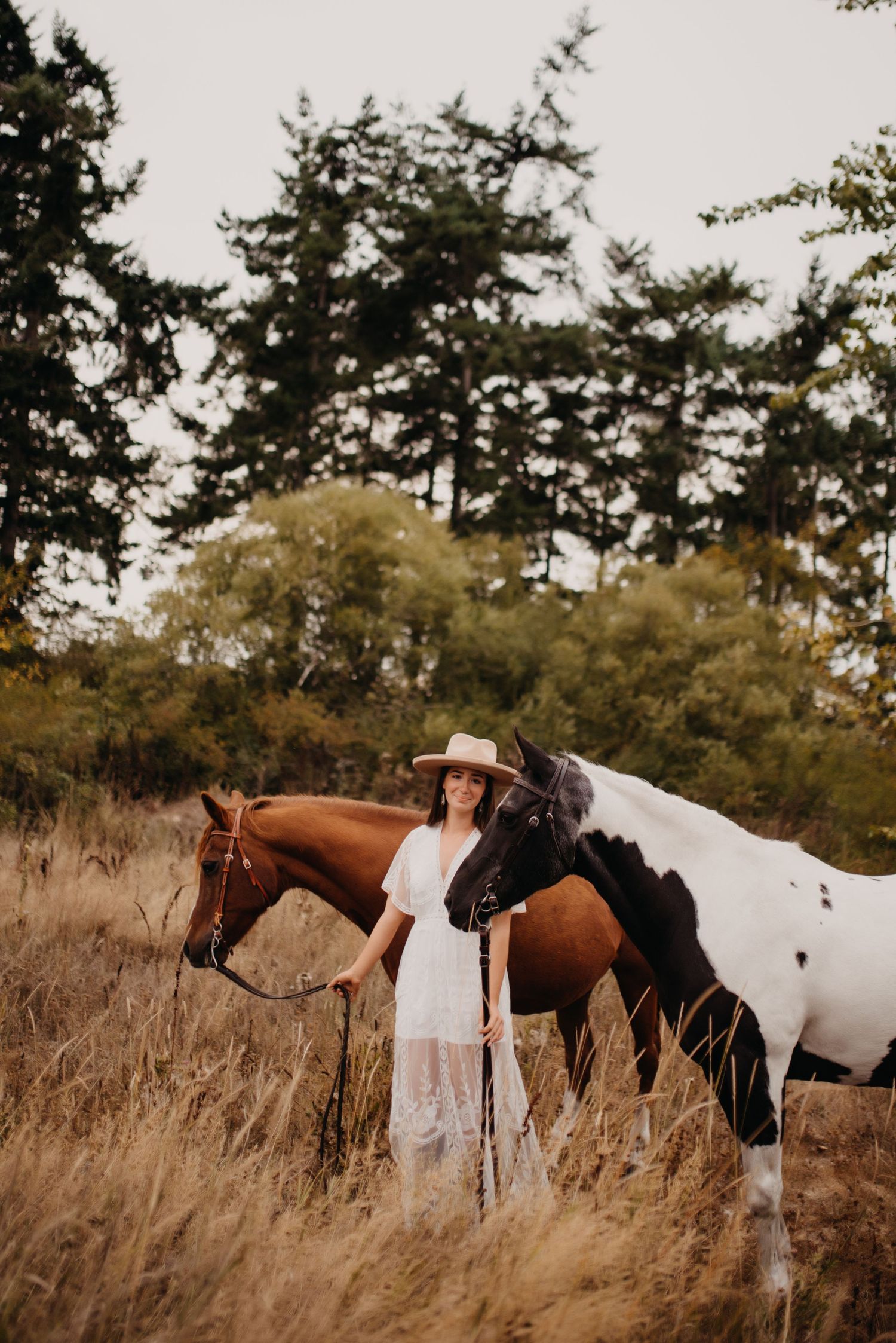 Young woman in white lace dress and hat standing with two horses in golden meadow.