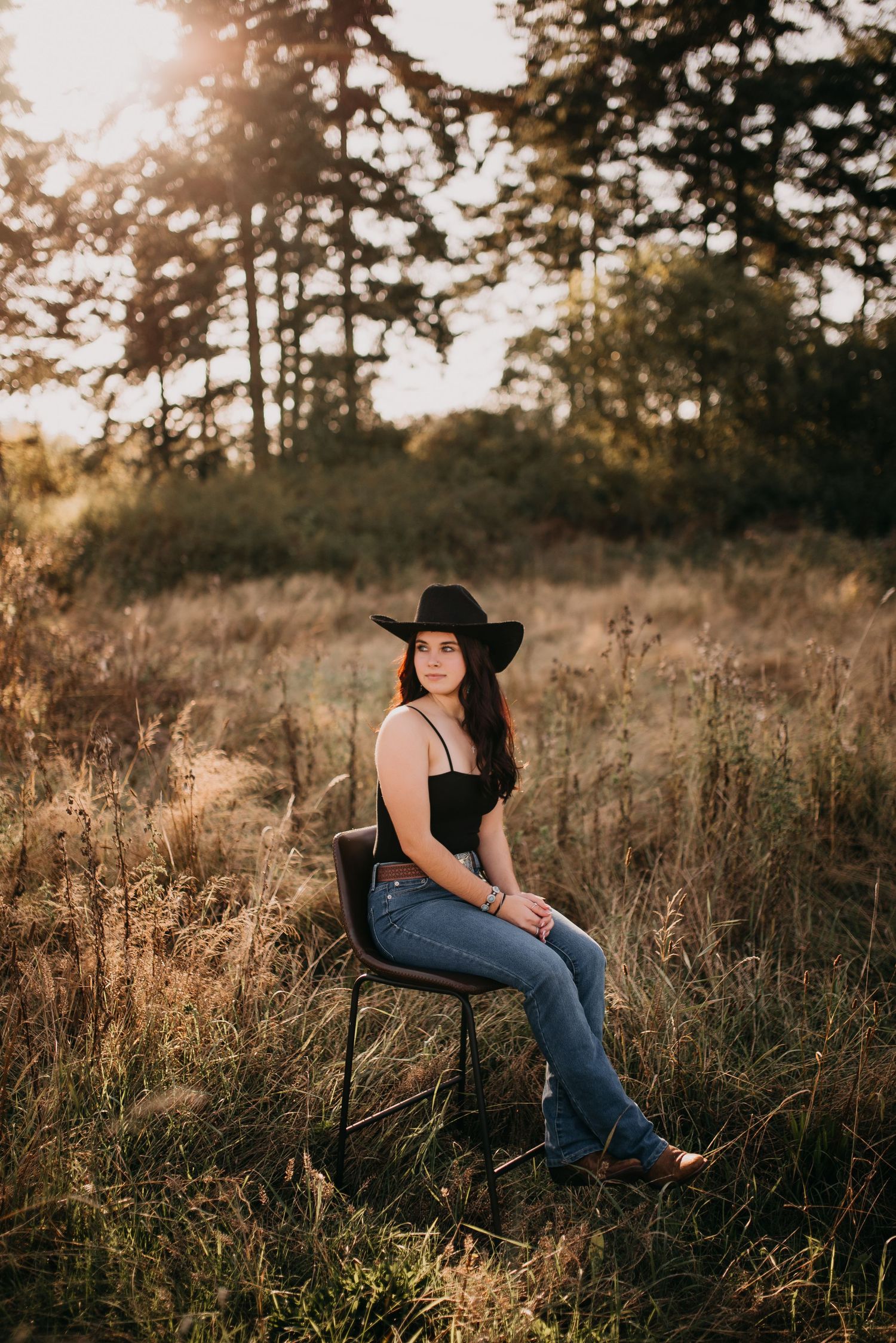 Brunette teen girl in black hat and jeans sitting on chair in golden sunlit meadow at sunset.