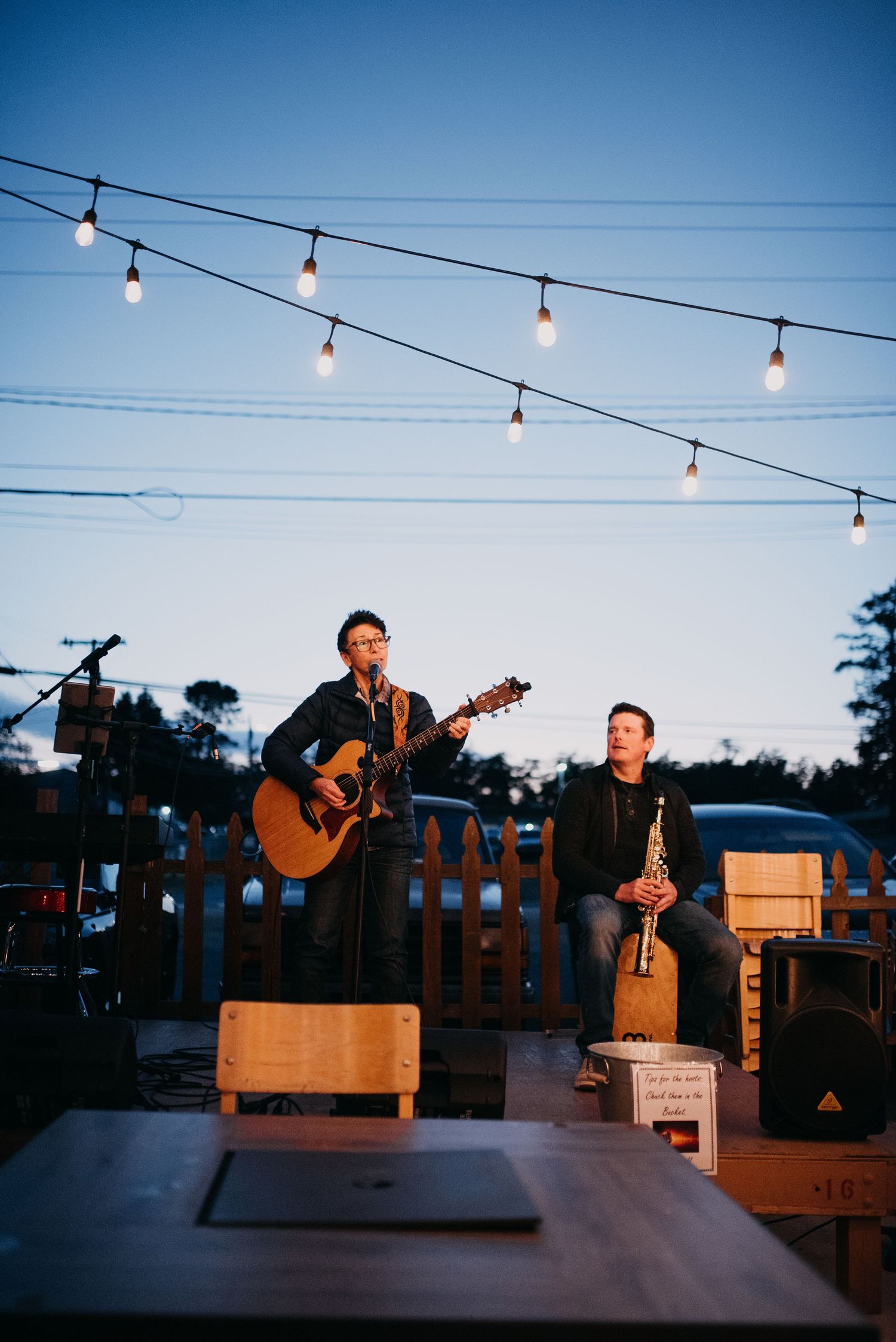Two musicians performing outdoors at dusk under string lights on a patio stage.