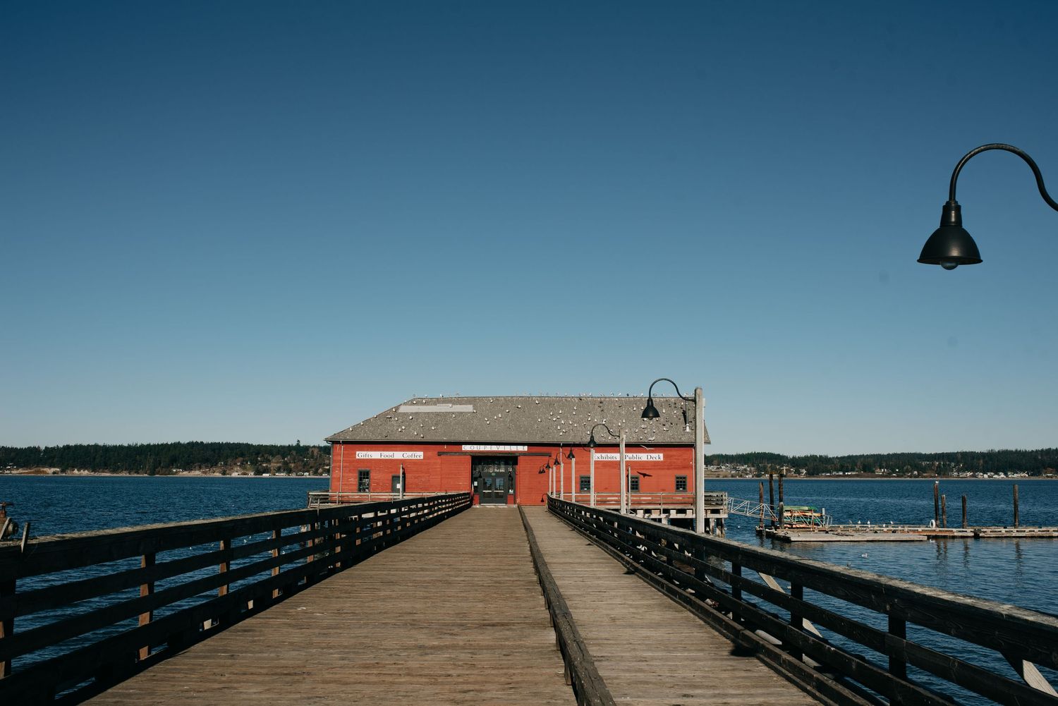 A red waterfront building on a wooden pier extending over calm blue waters under a clear sky.