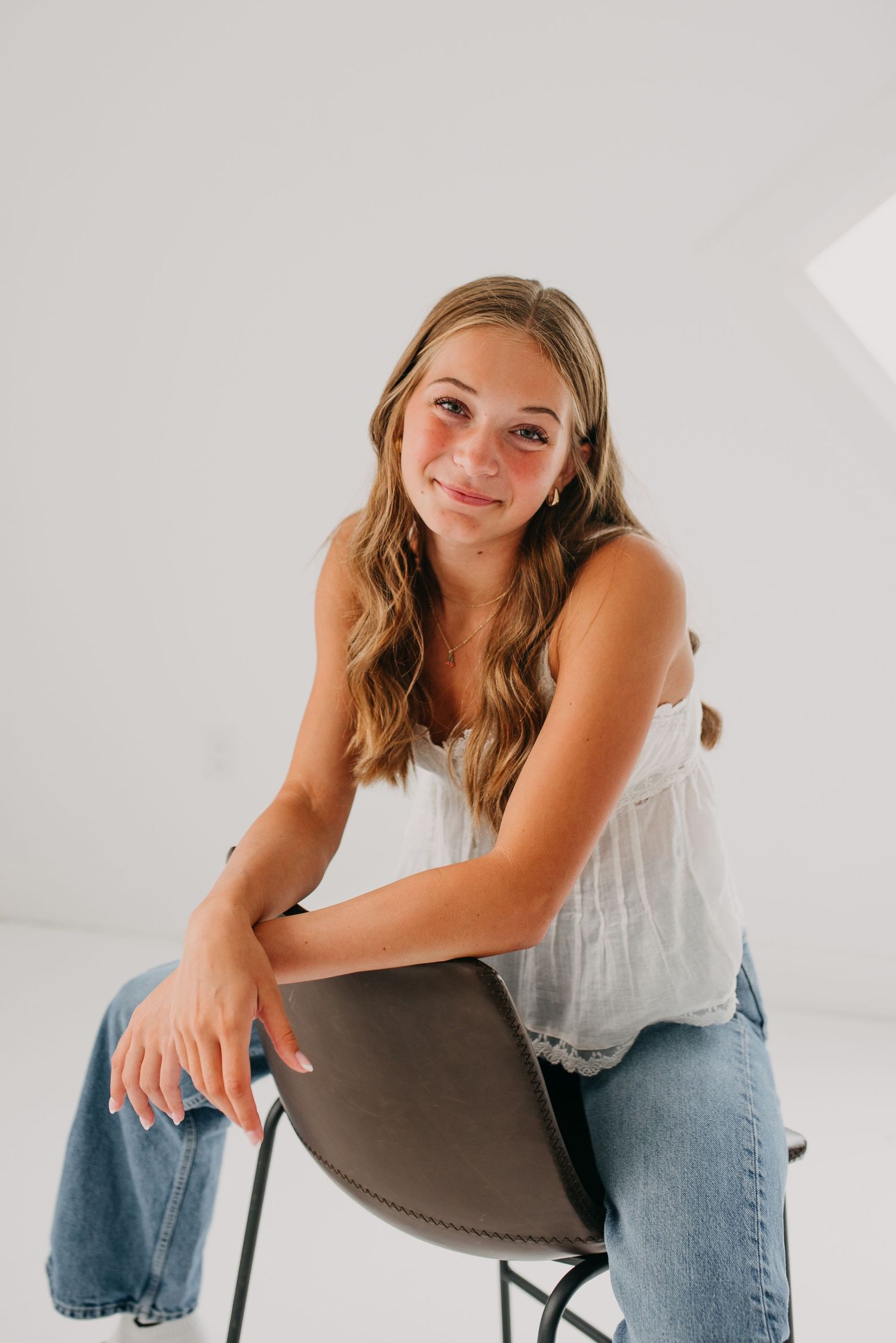 Teen girl with wavy hair in white tank top sitting on chair in bright white studio portrait.
