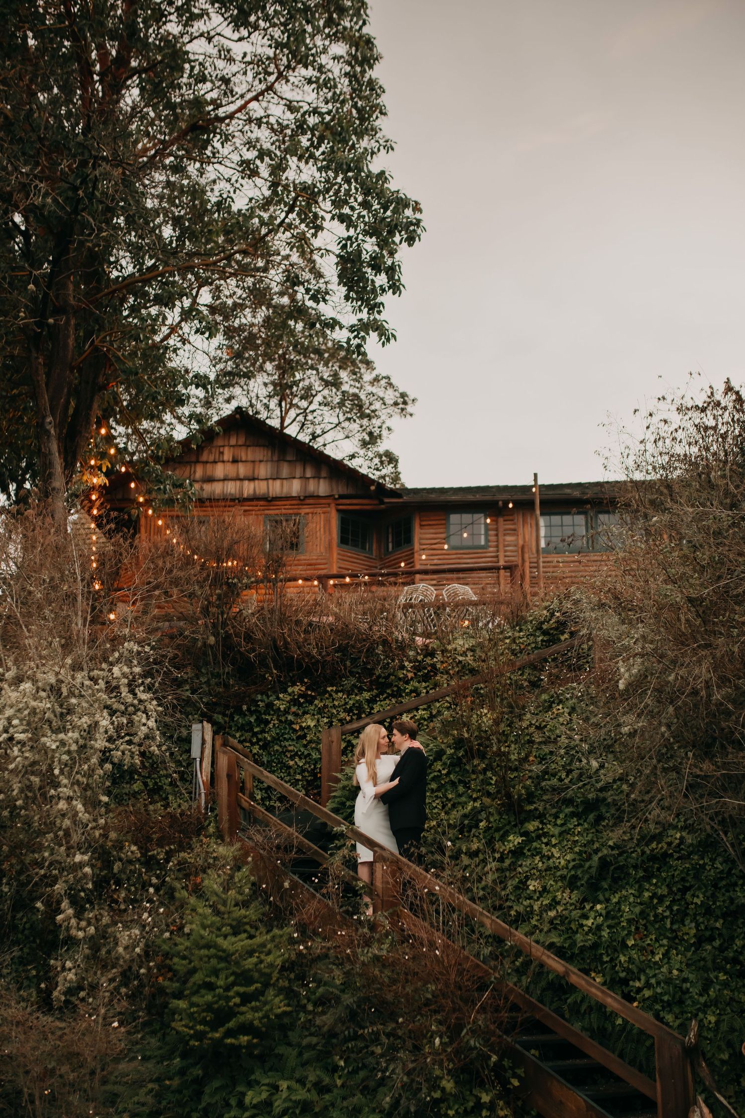 Bride and groom share a kiss on a wooden bridge surrounded by lush greenery and string lights.
