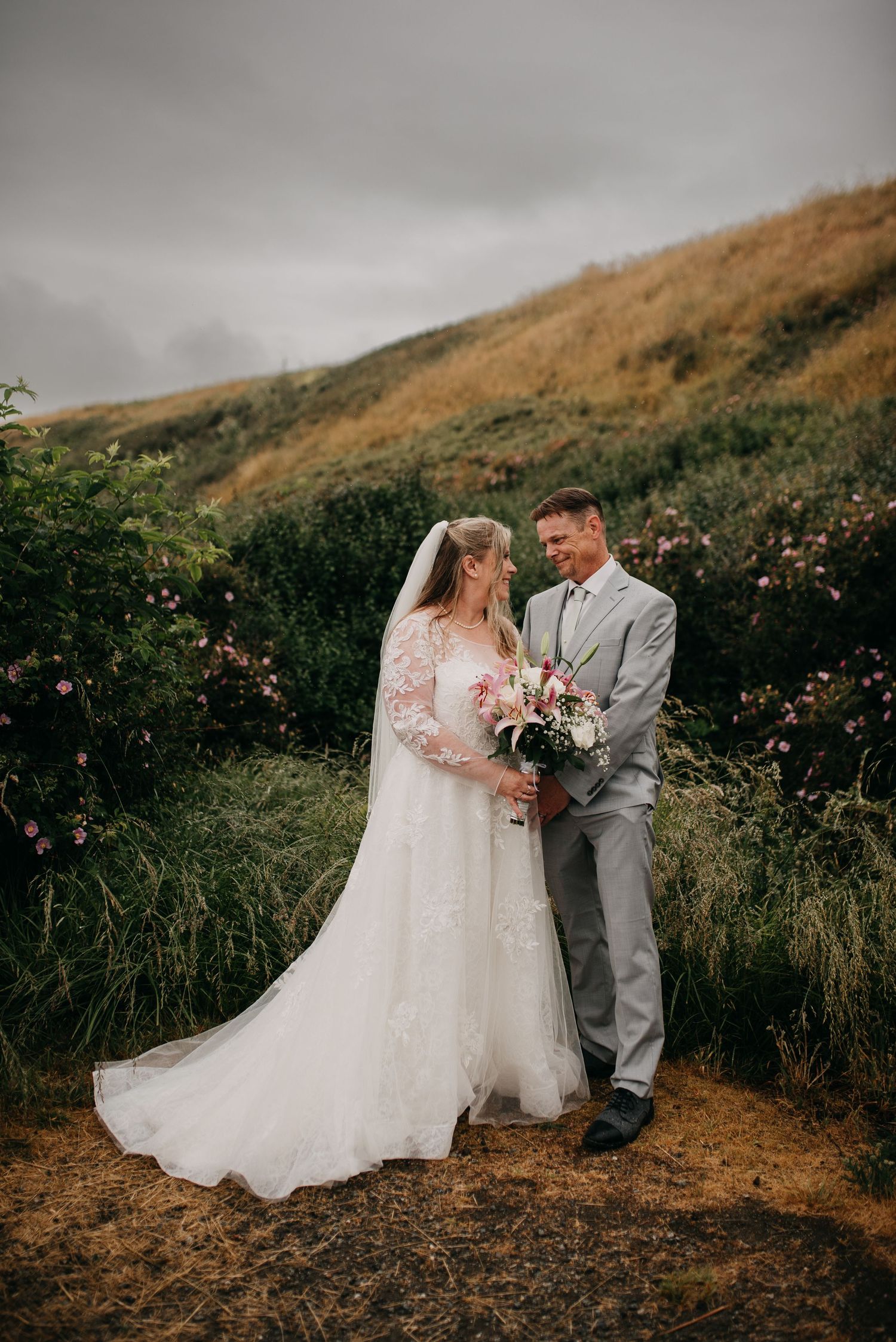 Bride in lace gown with veil and groom in gray suit pose with pink bouquet on a hillside meadow.