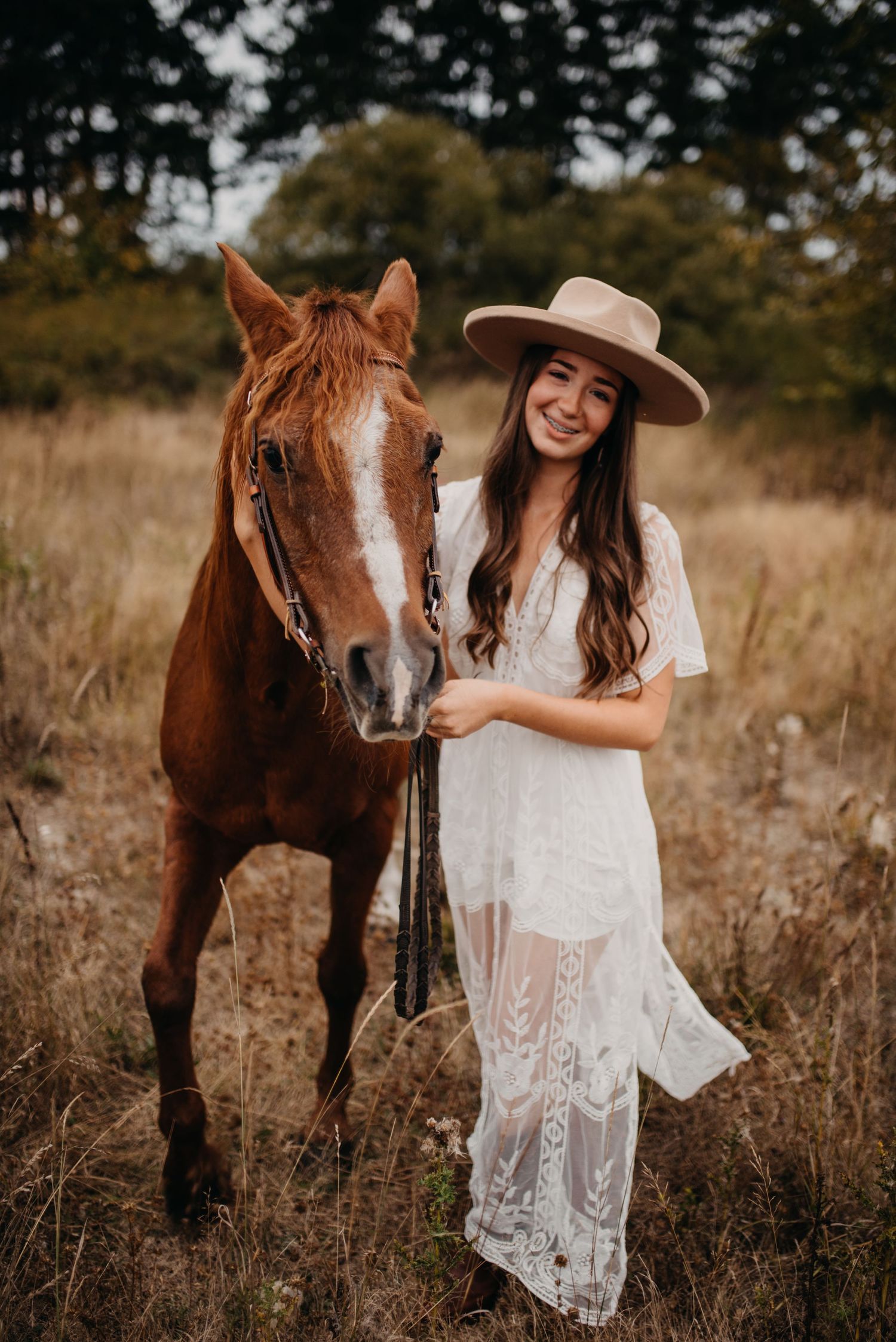 Smiling young woman in white lace dress and wide-brim hat posing with chestnut horse outdoors.