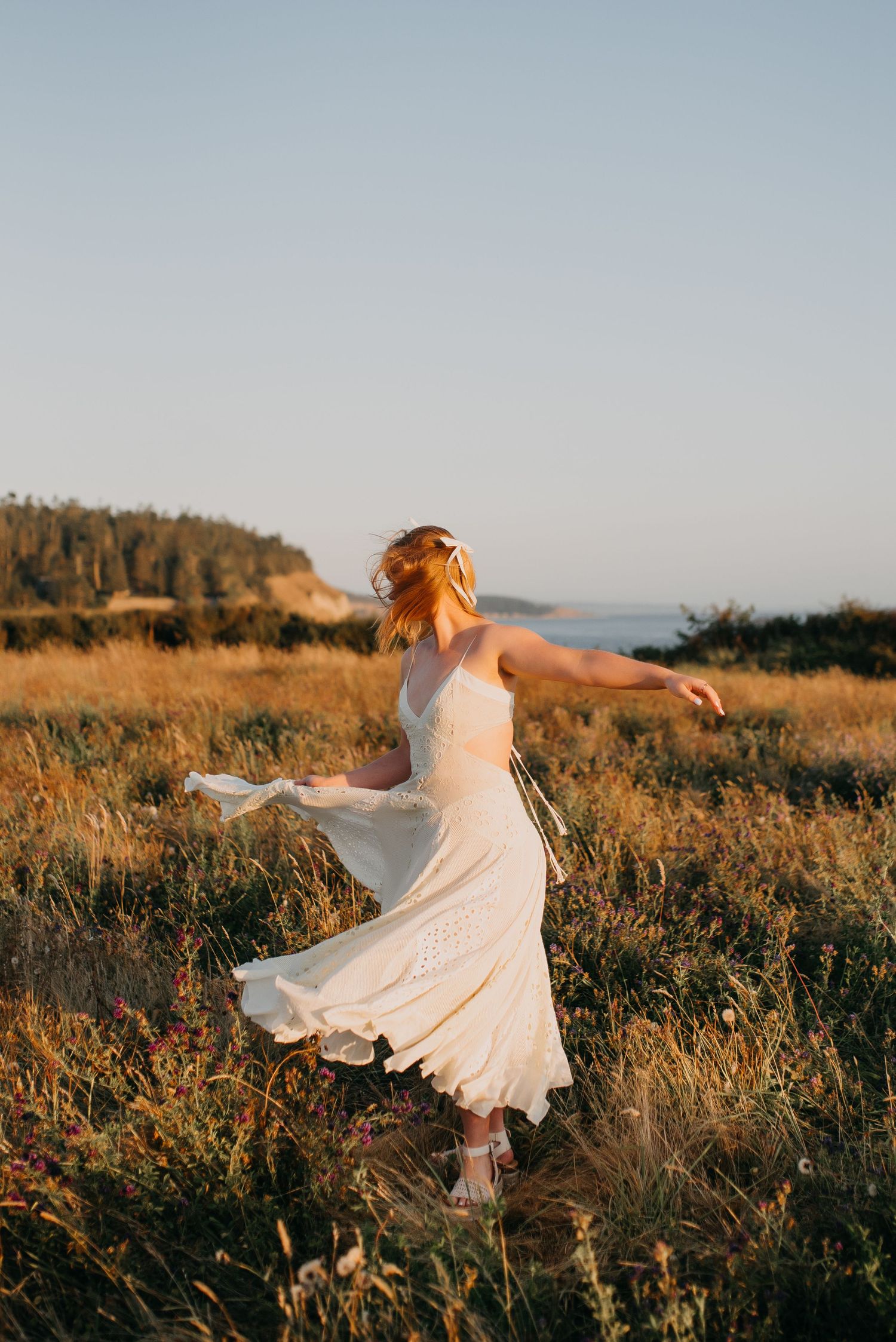 Woman in flowing white lace dress twirling in a golden meadow near the ocean at sunset.