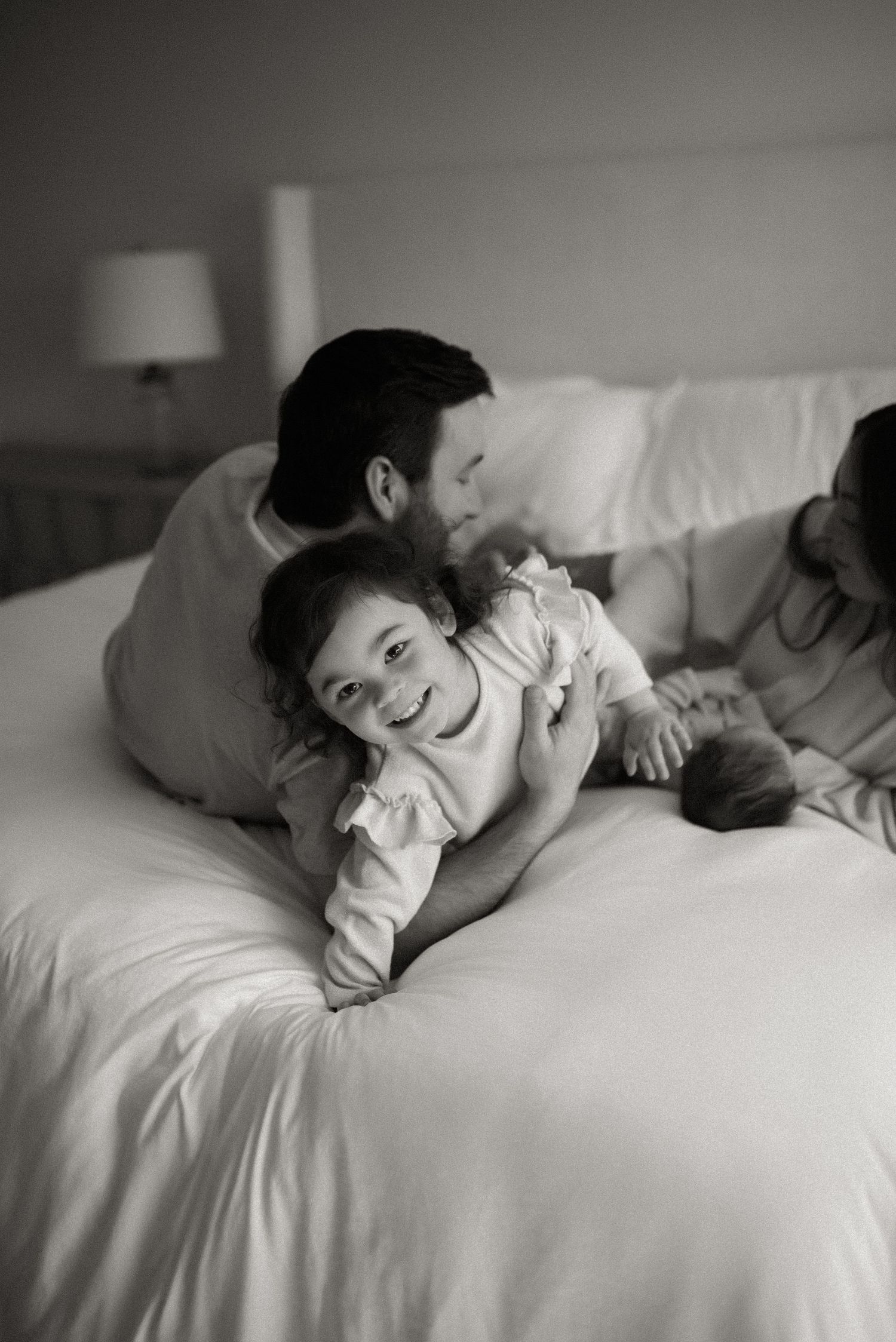 Black and white photo of father cuddling laughing toddler on white bed in cozy bedroom setting.