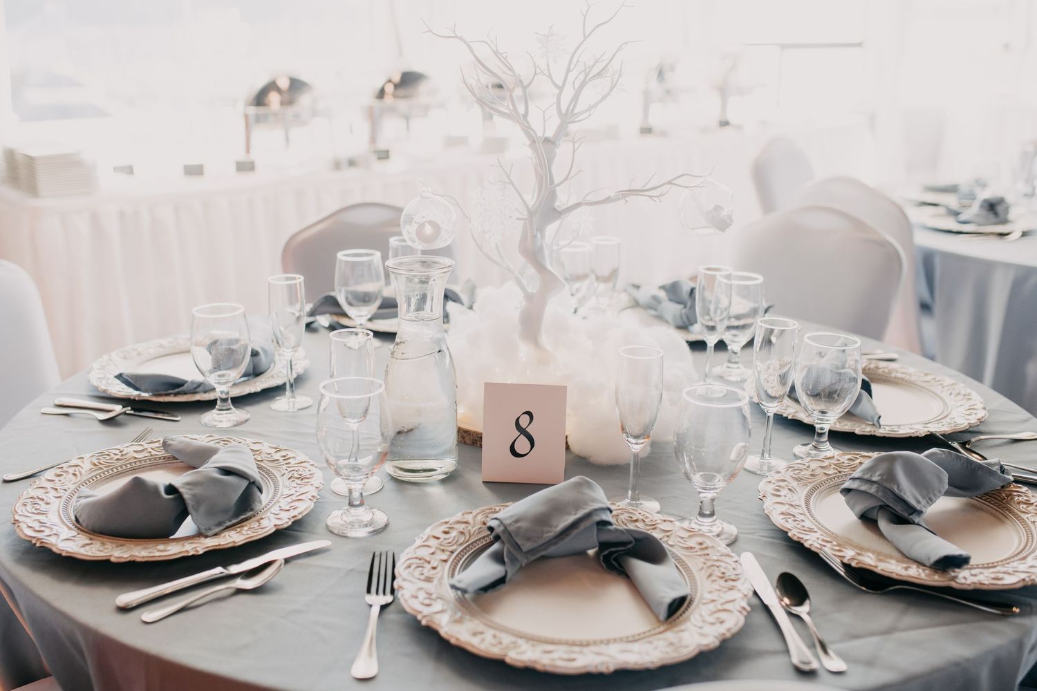 Round wedding reception table set with gray napkins, ornate chargers, and a bare branch centerpiece.