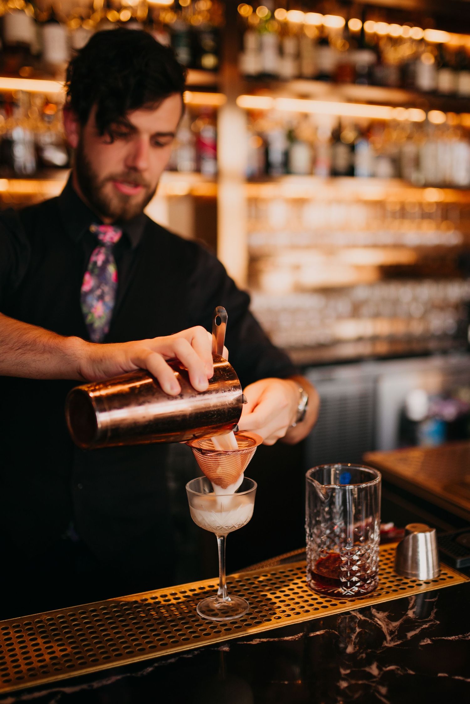 Bartender in black pouring a cocktail through a strainer into an elegant coupe glass at a bar.