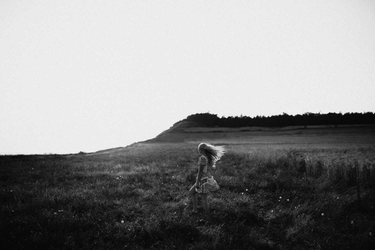 Black and white photo of a woman in a dress walking through an open field with a hill in the distance.