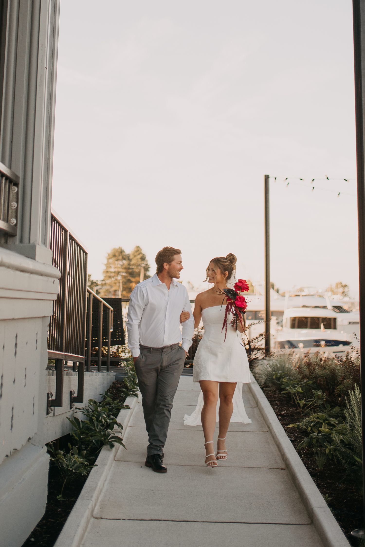Eloped couple walking along a marina pathway, bride holding vibrant red bouquet in mini white dress.