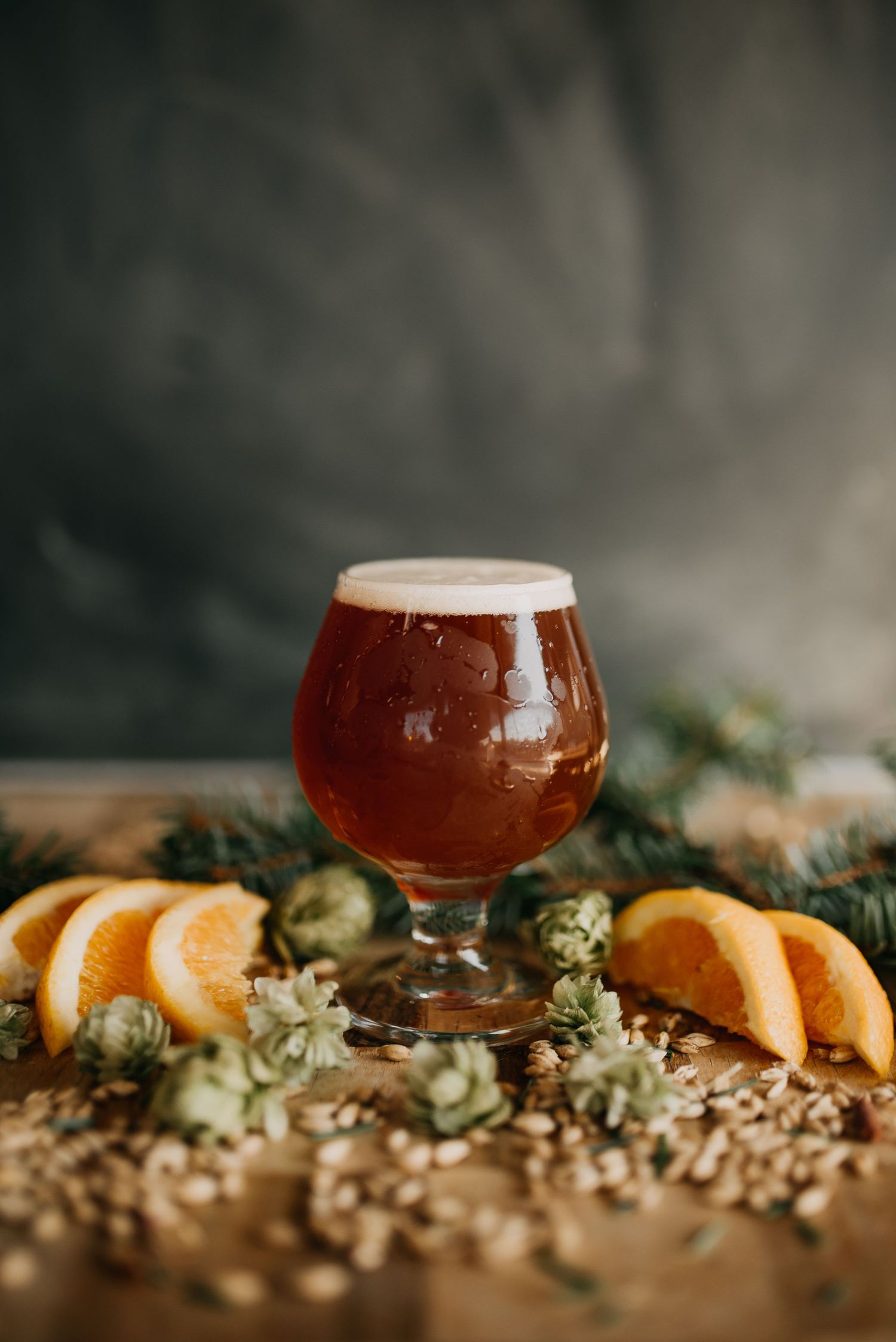 Amber craft beer in a tulip glass surrounded by orange slices, hops, and grain on a rustic wooden surface.