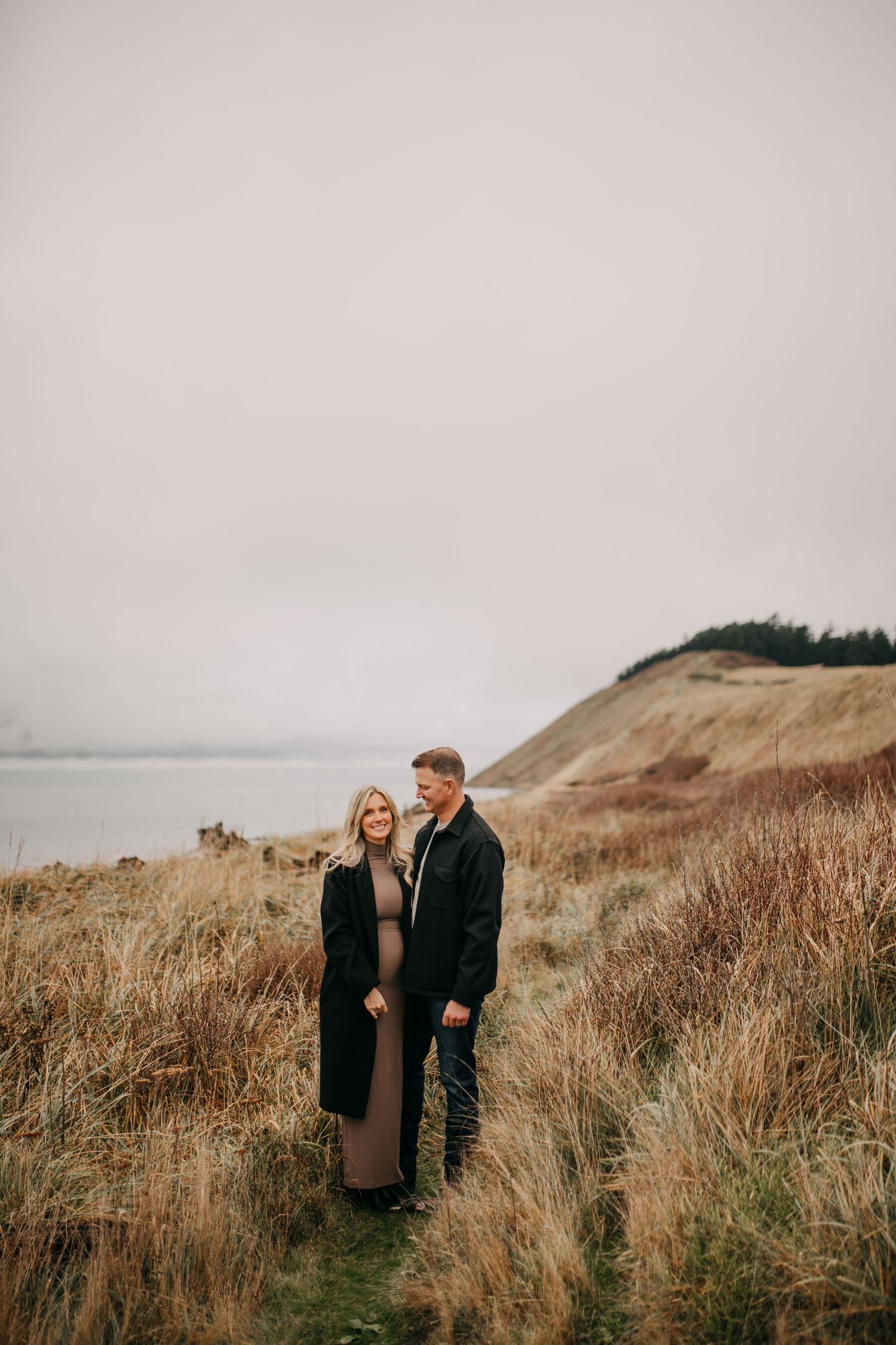 Couple stands together in coastal tall grass field on overcast day near water.