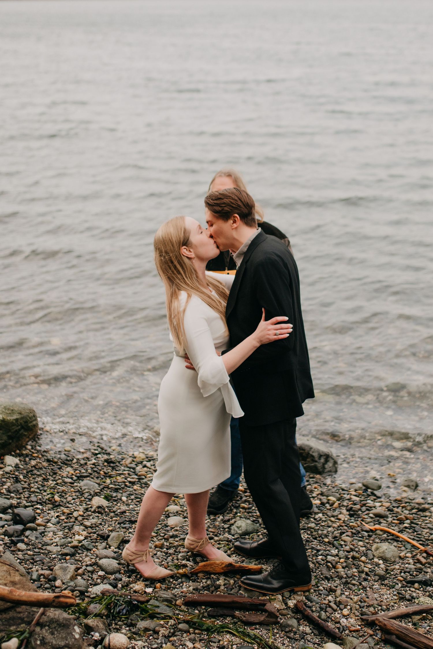 Couple sharing a romantic kiss on a rocky beach shoreline with calm ocean waters in the background.