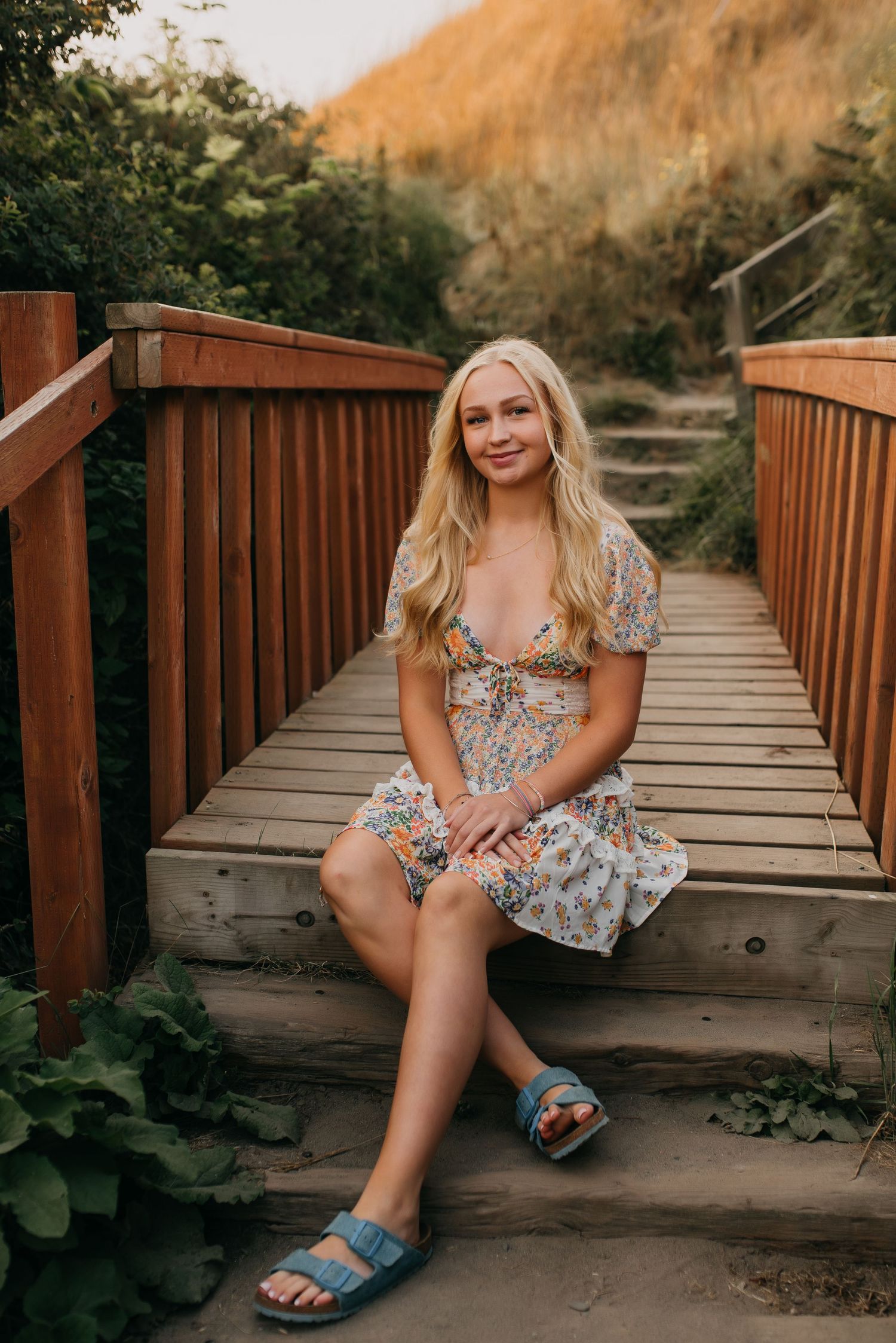 Smiling blonde woman in floral mini dress sits on a wooden boardwalk bridge surrounded by lush greenery.