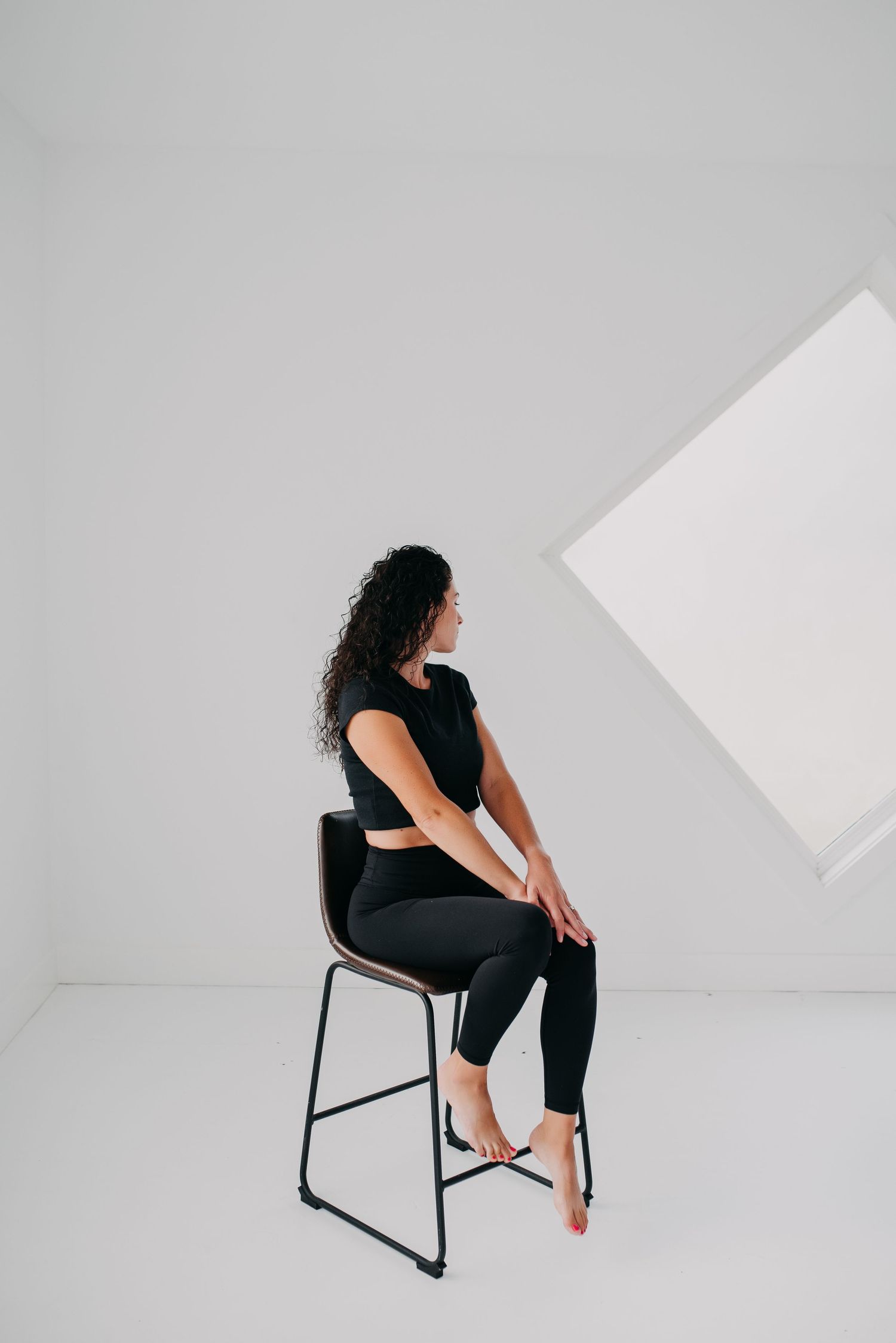Woman in black athletic wear sitting on a modern chair in a minimalist white studio space.