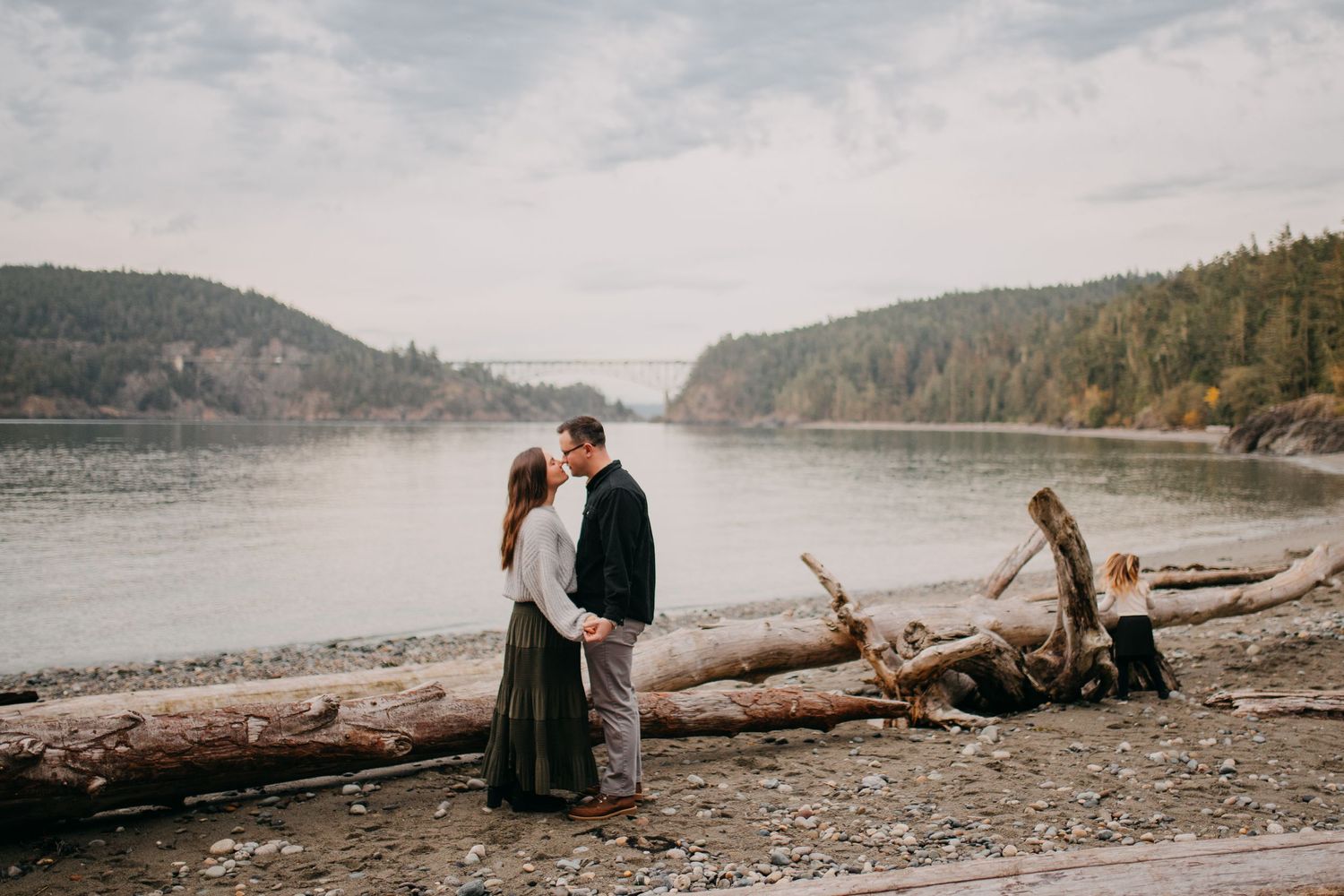Couple sharing romantic kiss near driftwood logs with scenic bridge and forested river in background.