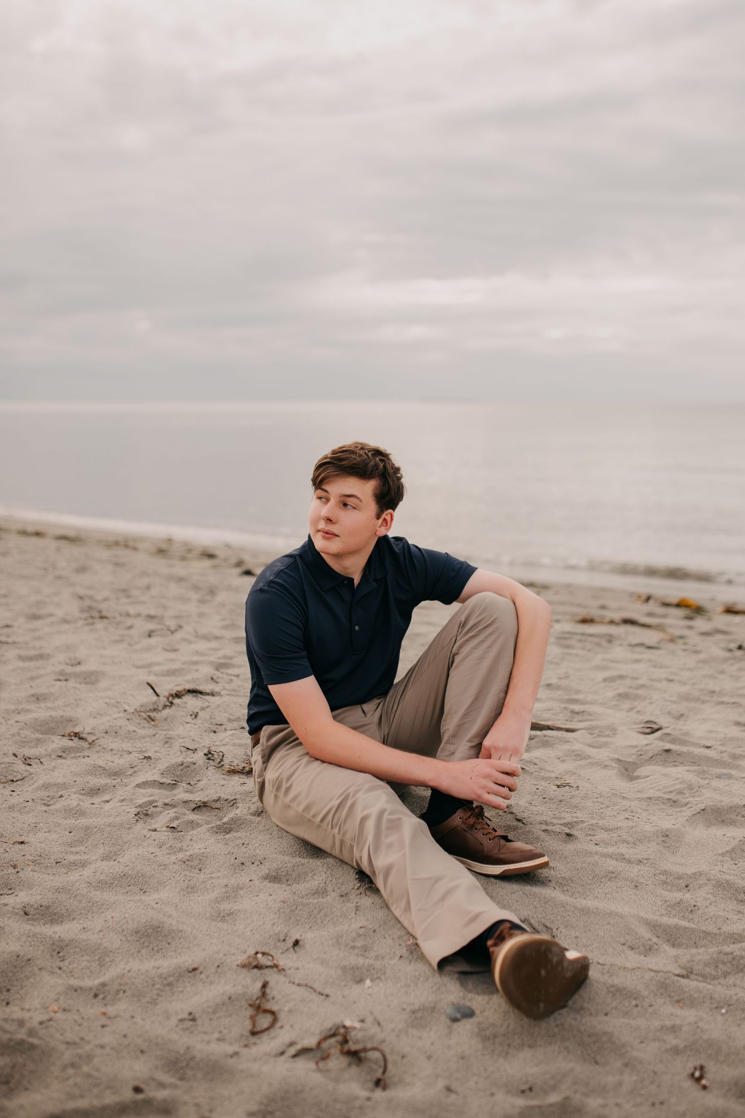 Young man in navy polo and khakis sits relaxed on sandy beach with cloudy sky.