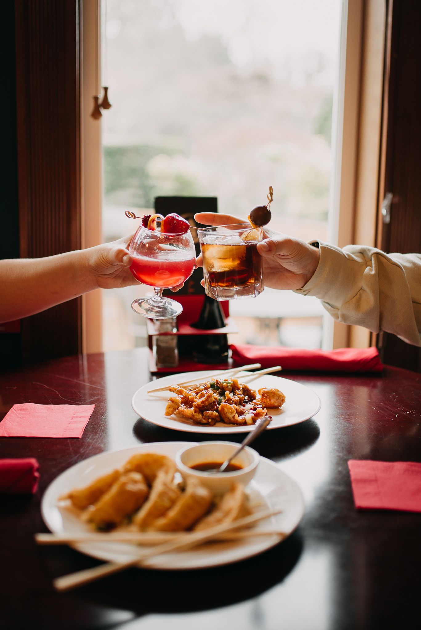 Two people toasting cocktails over plates of dumplings and orange chicken at a restaurant table.