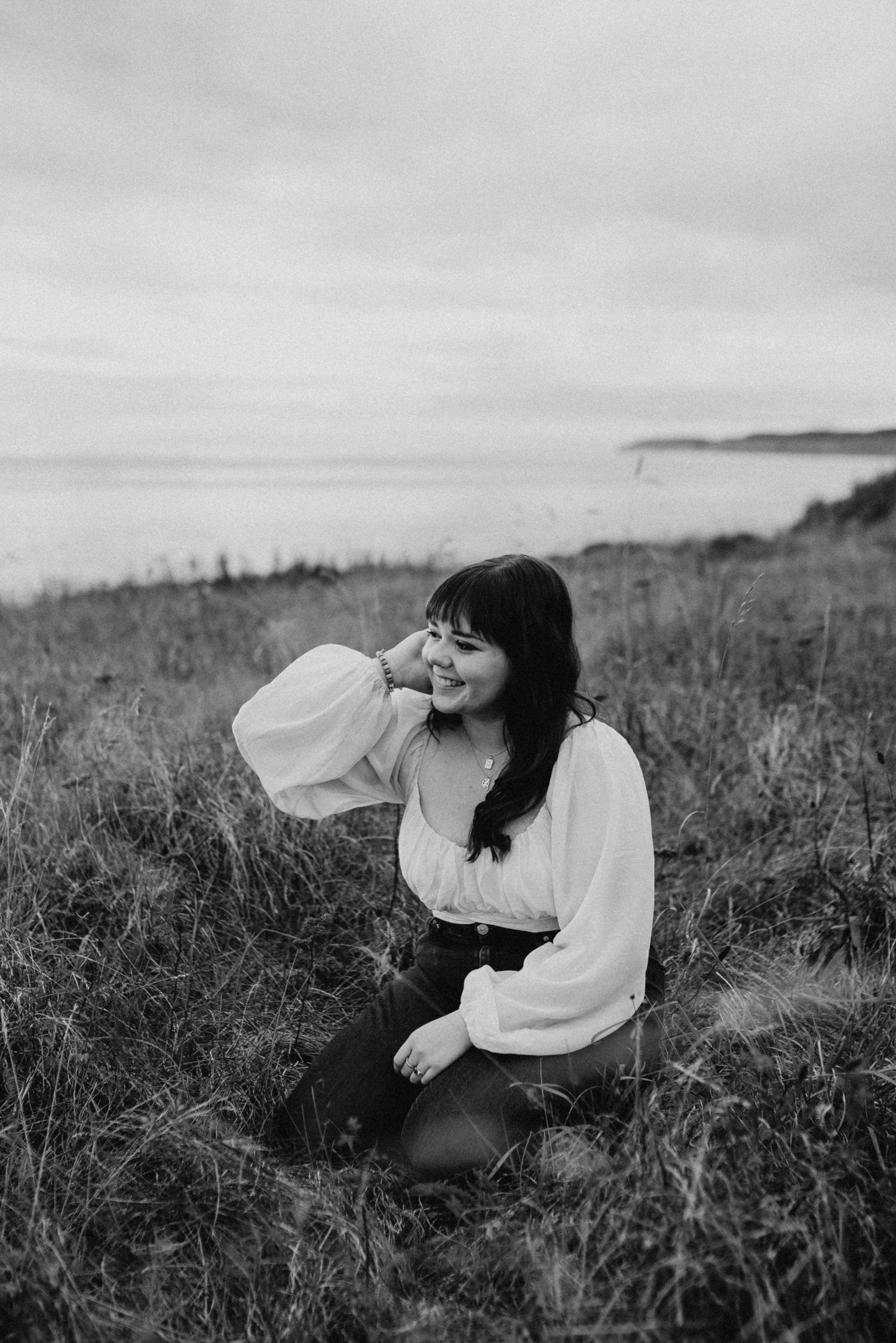 Young woman sitting in coastal grass field smiling, wearing white blouse, black and white portrait photography.