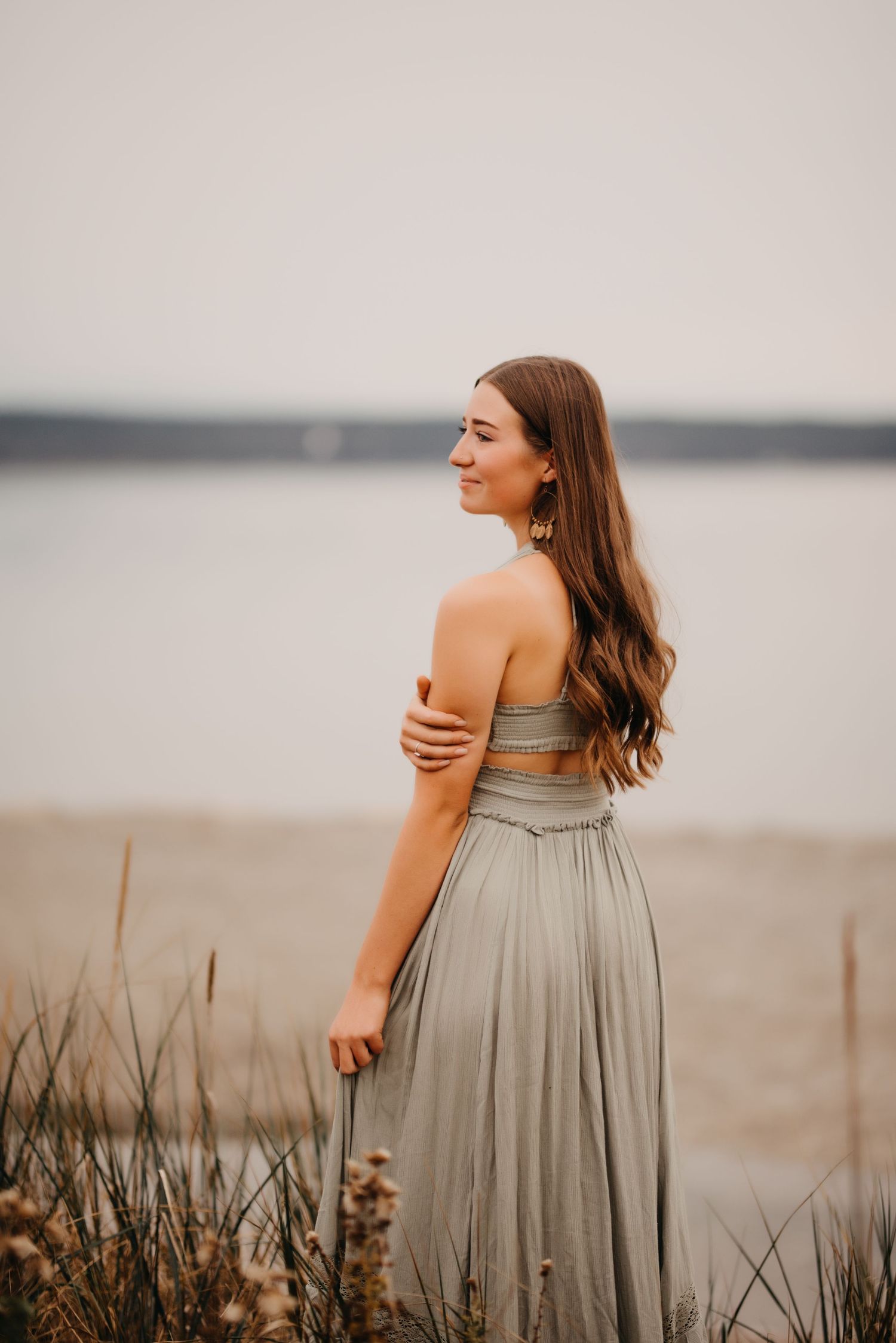 Young woman in sage green strapless dress stands by waterfront, looking over her shoulder at dusk.