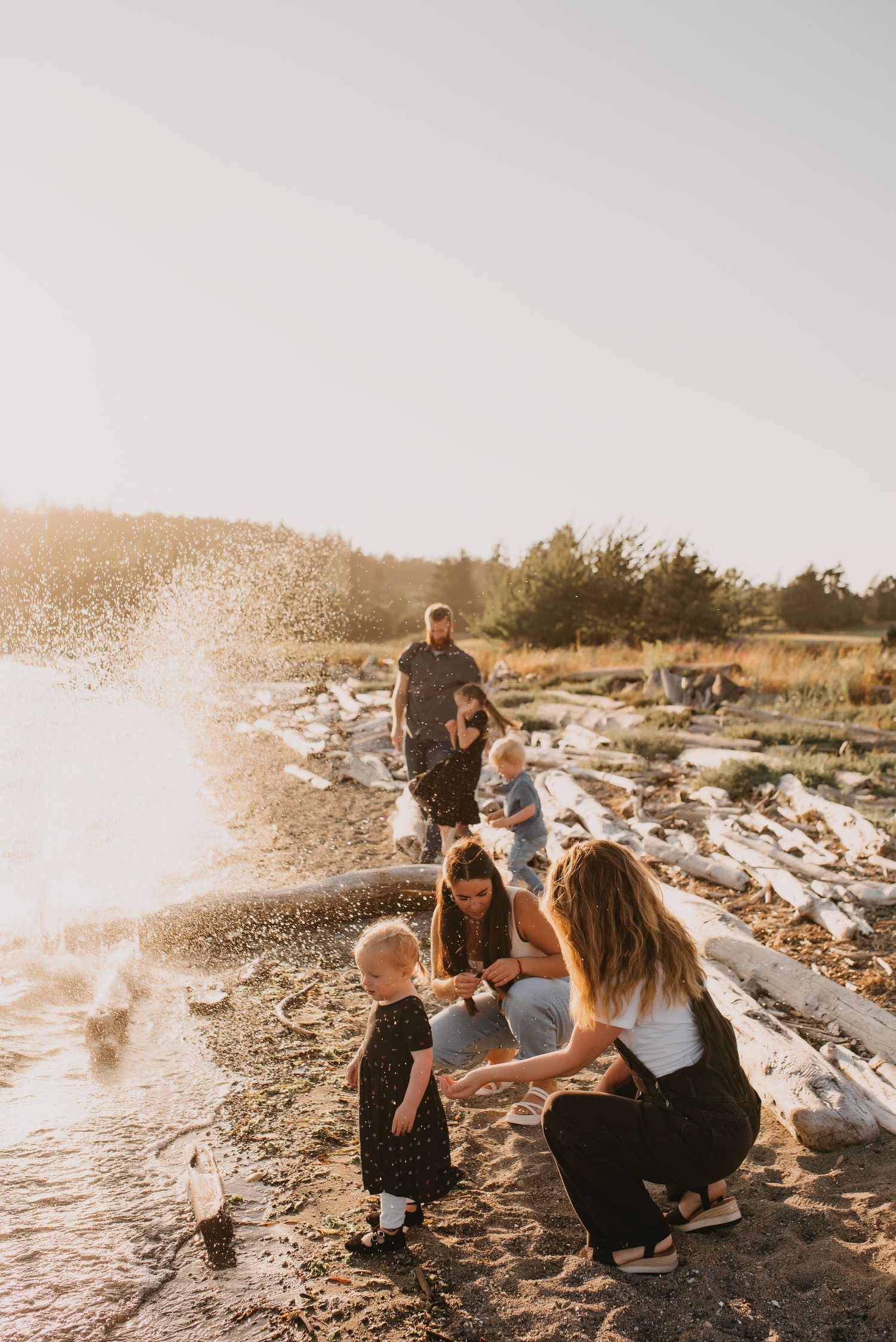 Family enjoying beach at sunset with children playing near water and driftwood logs.