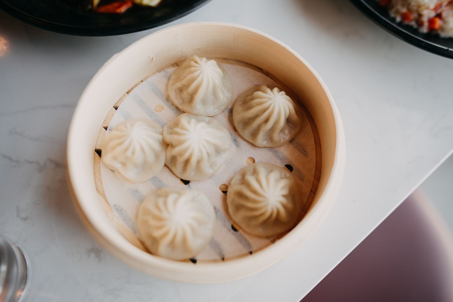 Close-up of six delicate xiaolongbao soup dumplings in bamboo steamer basket on white table.