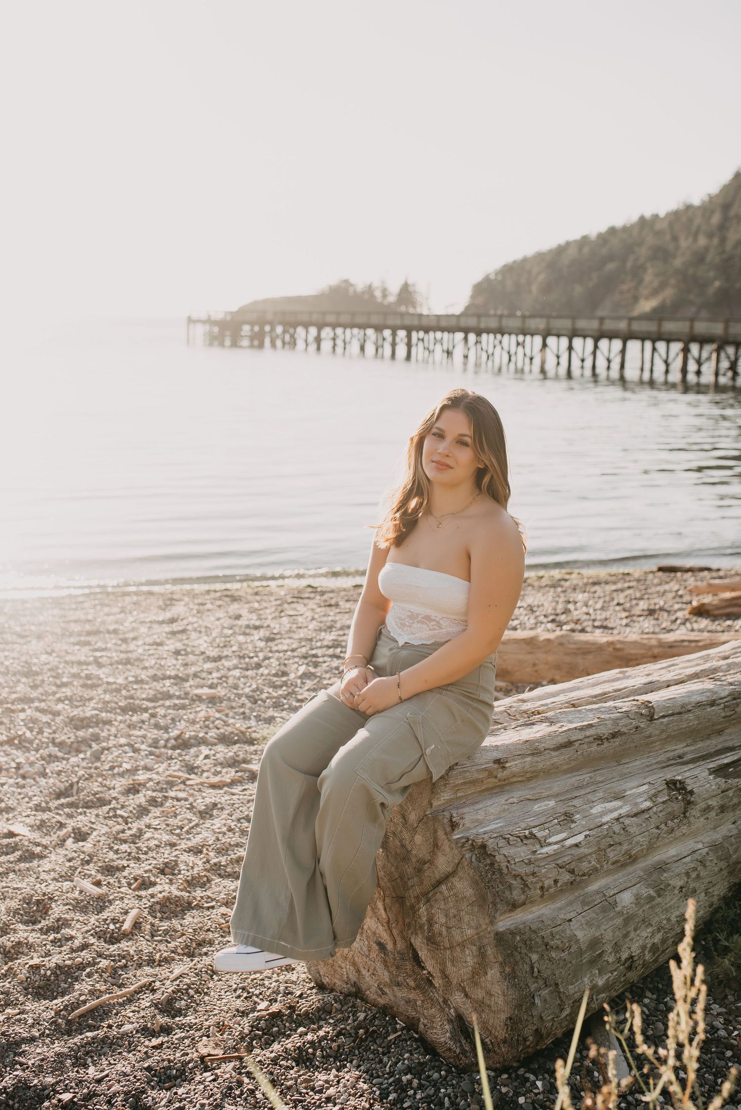 Teen girl in white tube top and sage green pants sitting on driftwood log at beach during golden hour.