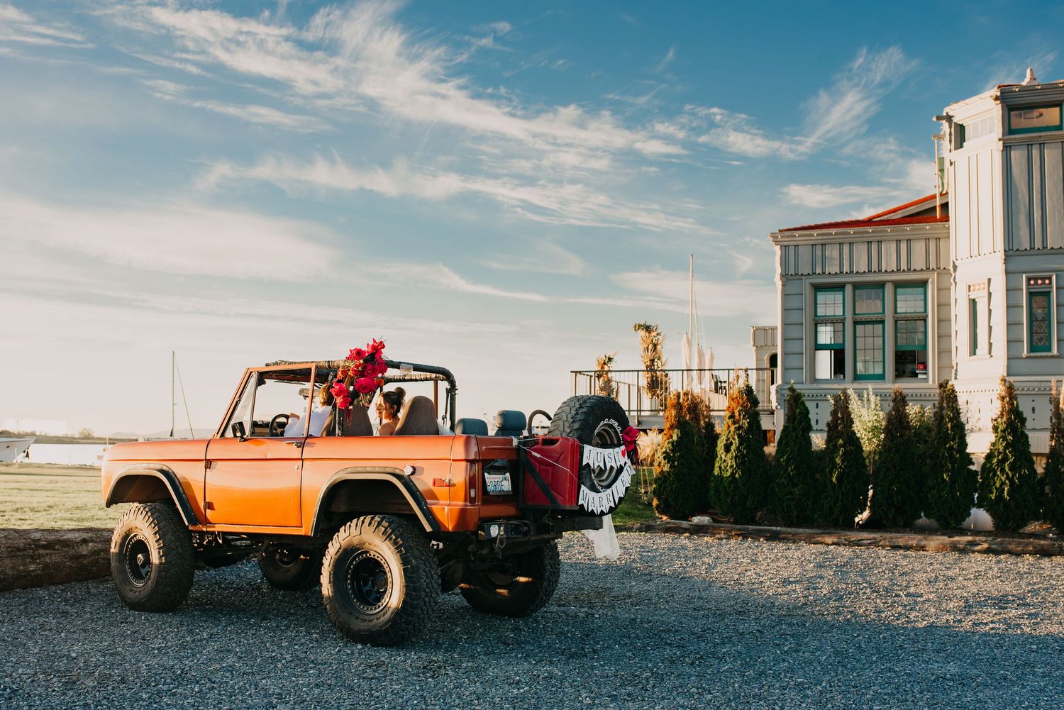 Vintage orange open-top Bronco parked on gravel beside tall hedges and an elegant building under blue sky.
