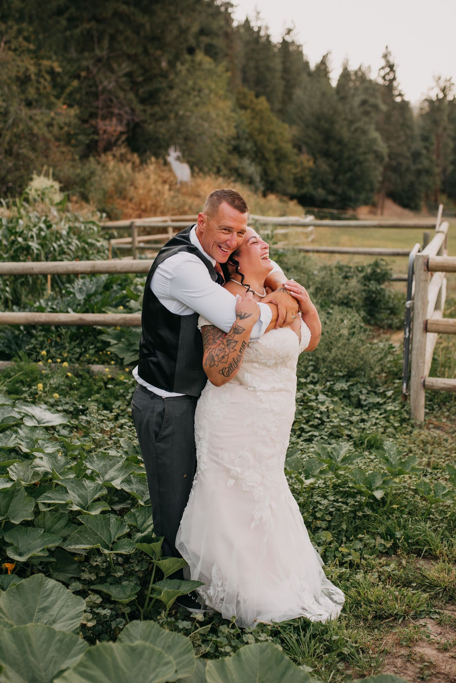 Tattooed groom embraces smiling bride in white lace dress beside a rustic wooden fence in a lush garden.