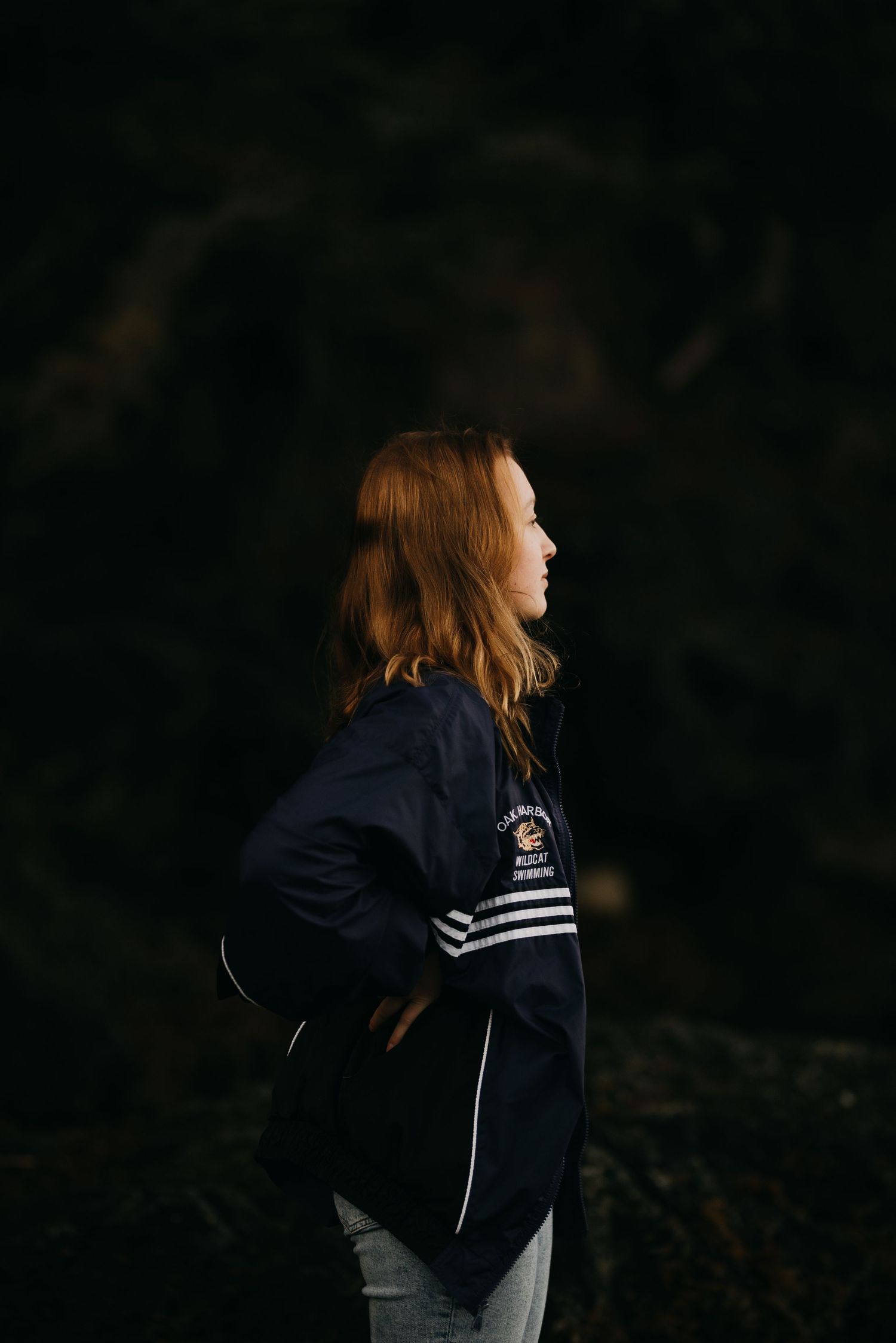 Young woman with red hair in navy varsity jacket standing in profile against dark background.