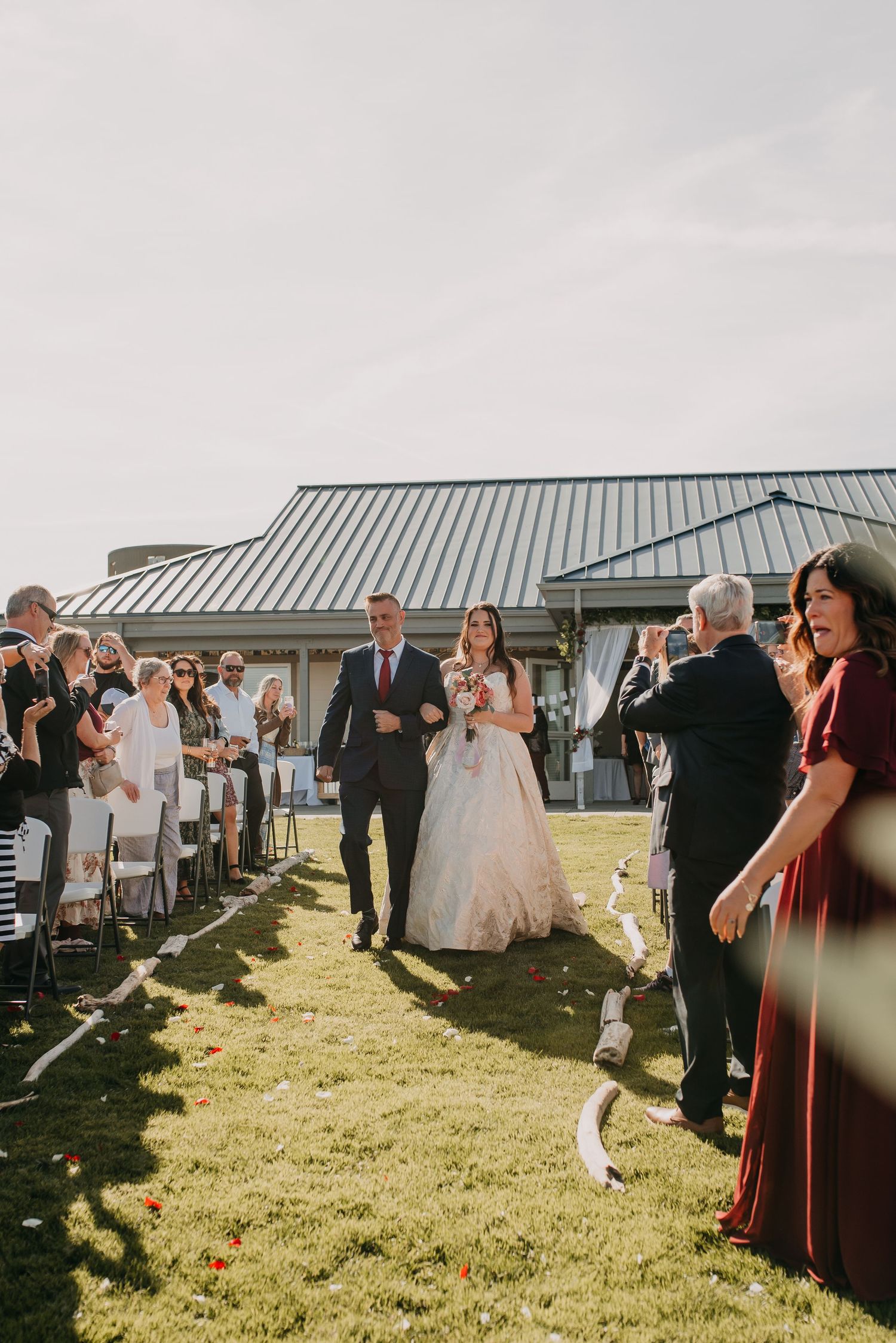 Bride and groom walk down outdoor wedding aisle as guests celebrate with streamers on sunny day.