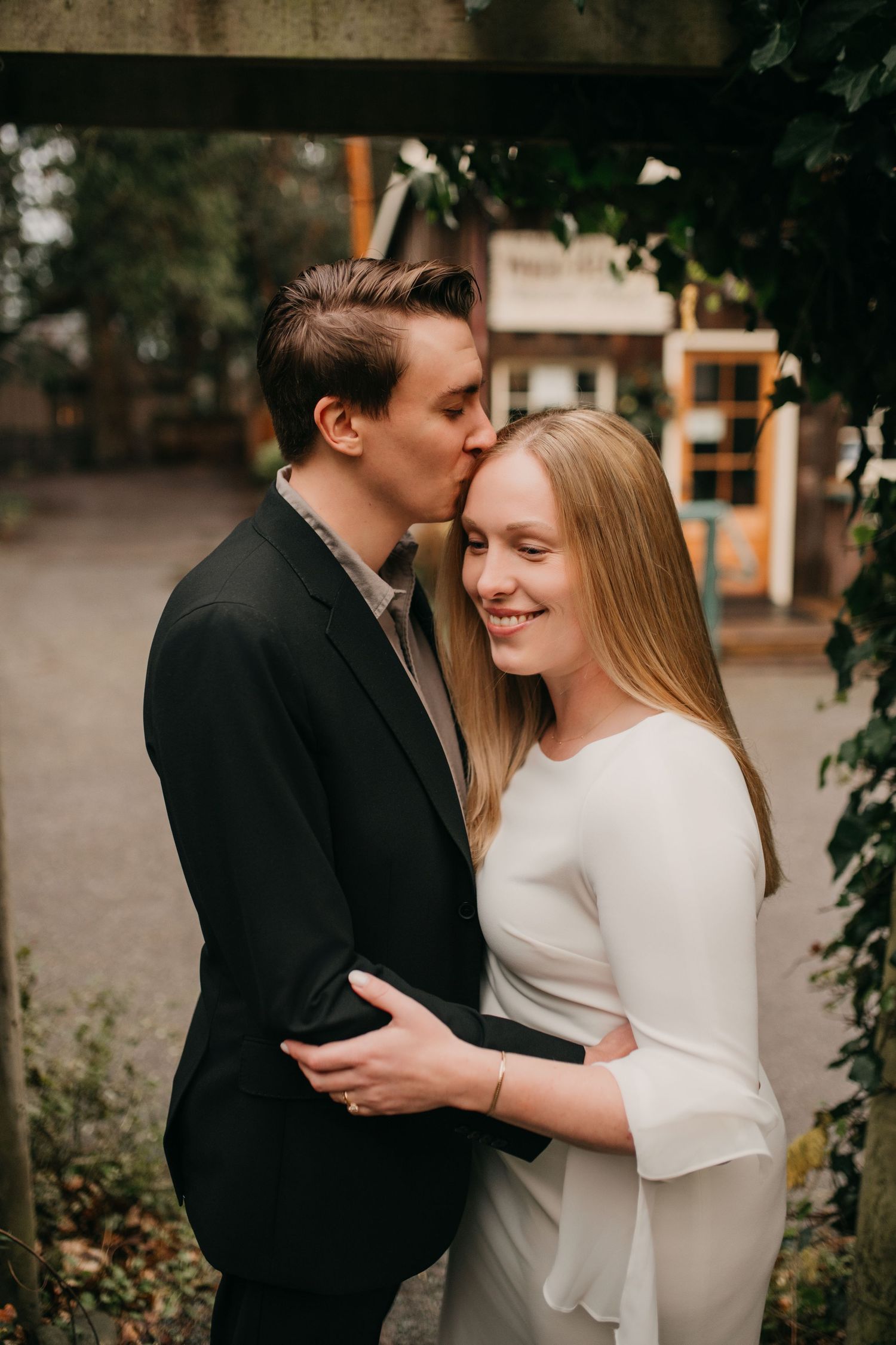 Groom kisses smiling bride on cheek during romantic outdoor engagement photo session.