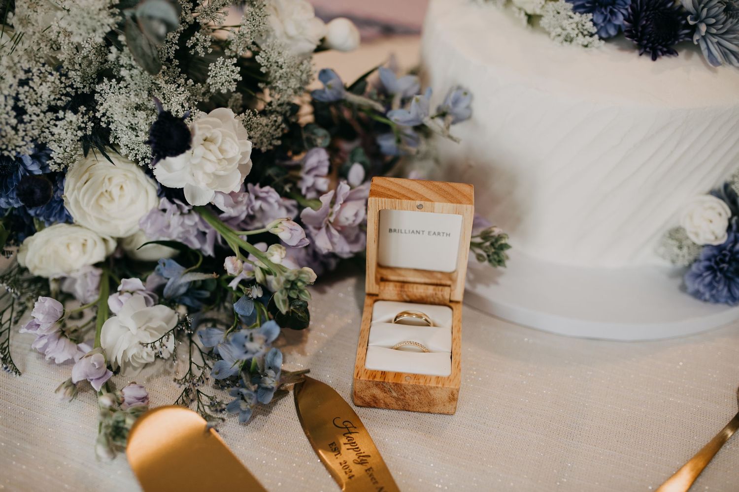 Gold wedding rings in wooden box displayed beside blue and white floral bouquet and wedding cake.