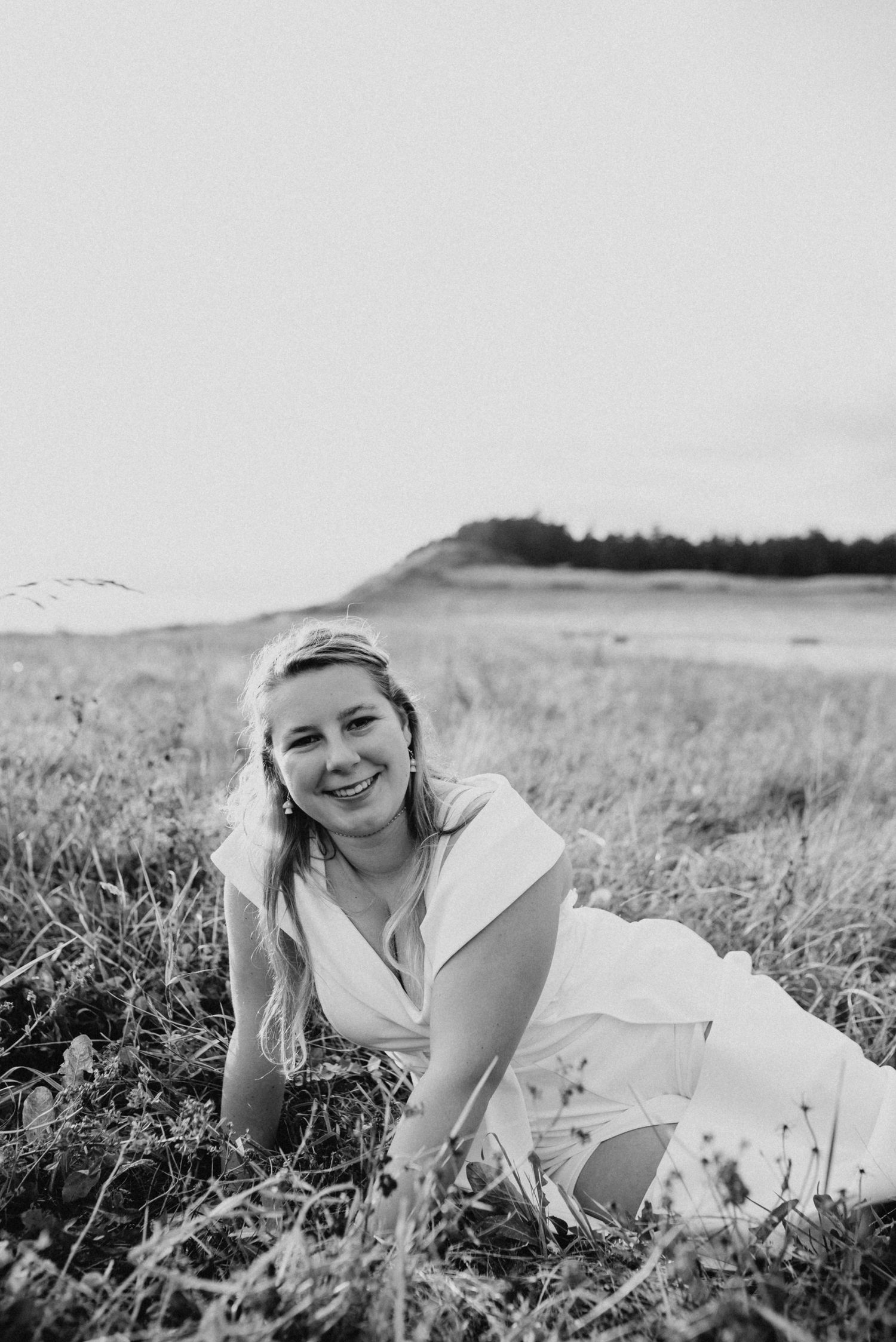 Smiling young woman in white dress lying in field of tall grass in black and white senior photo.