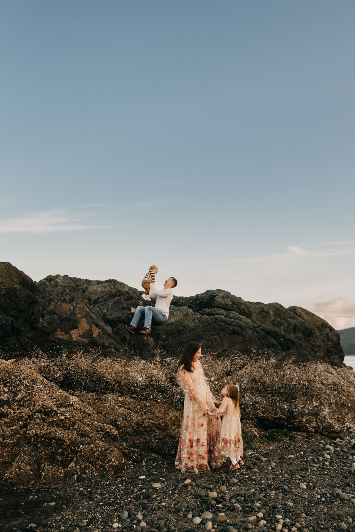 Father lifting baby on rocky coastal cliffs while mother and daughter watch below.