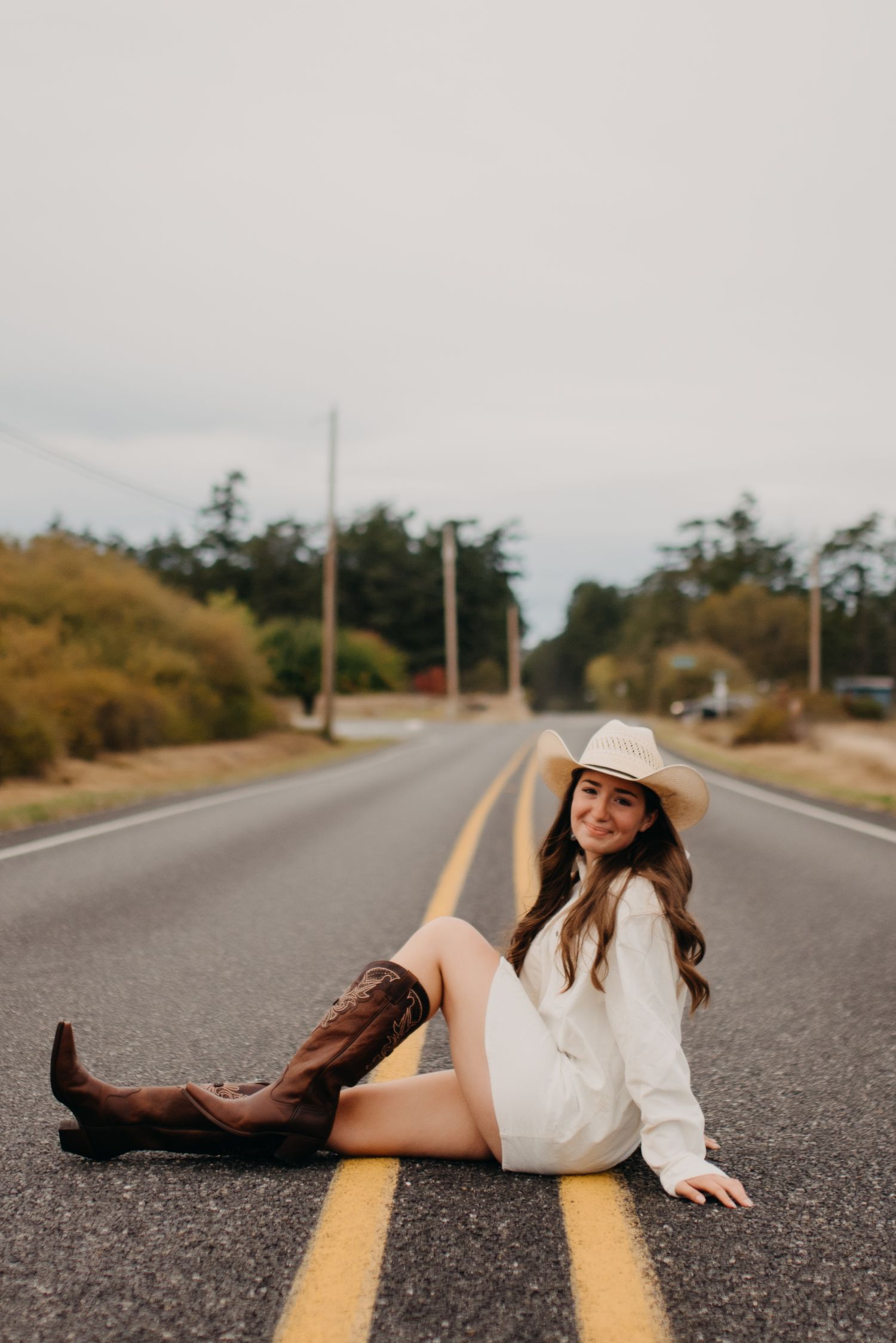 Young woman in white dress and cowboy hat sitting on road centerline with brown boots.