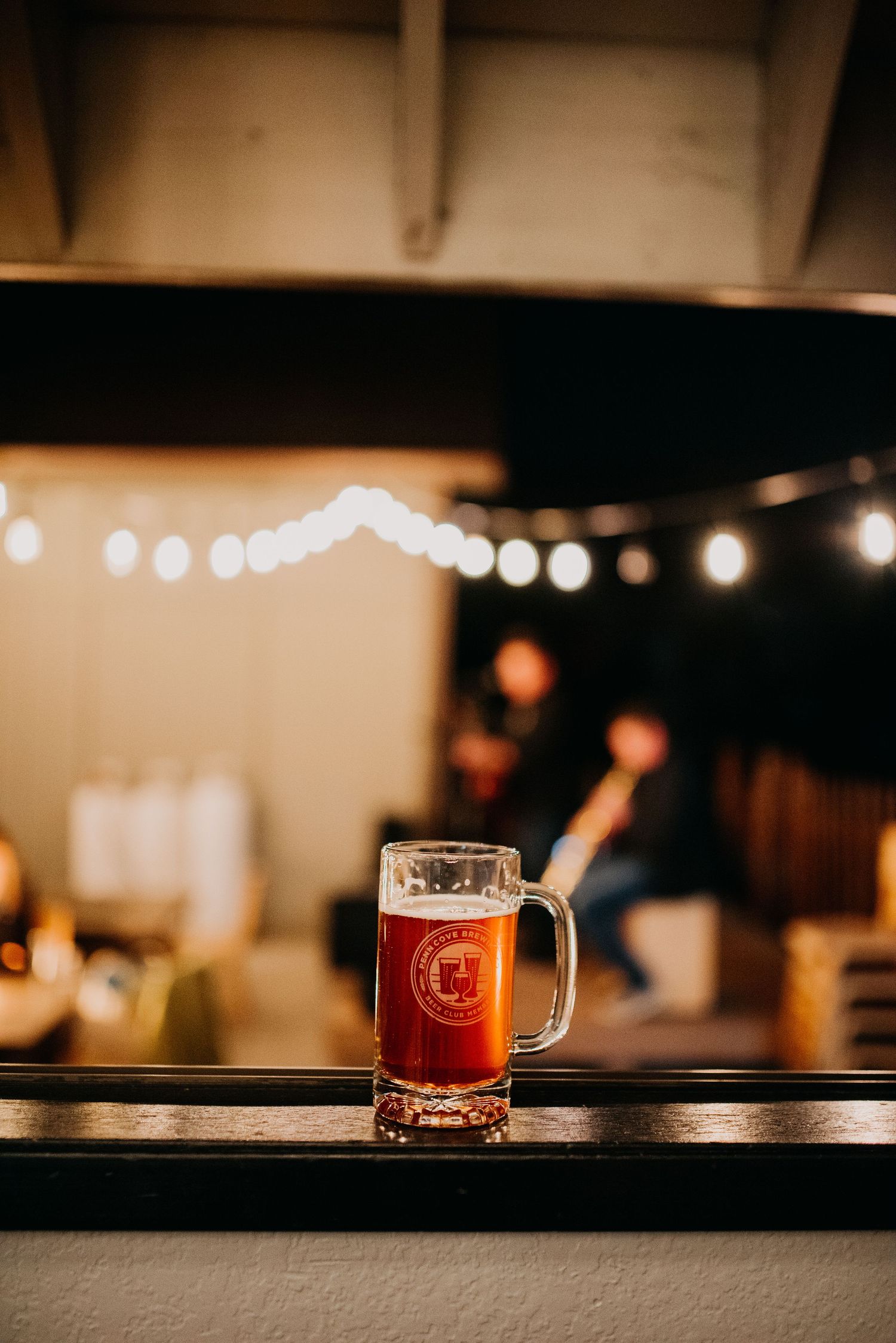 Branded beer mug filled with amber ale on a bar ledge with string lights.