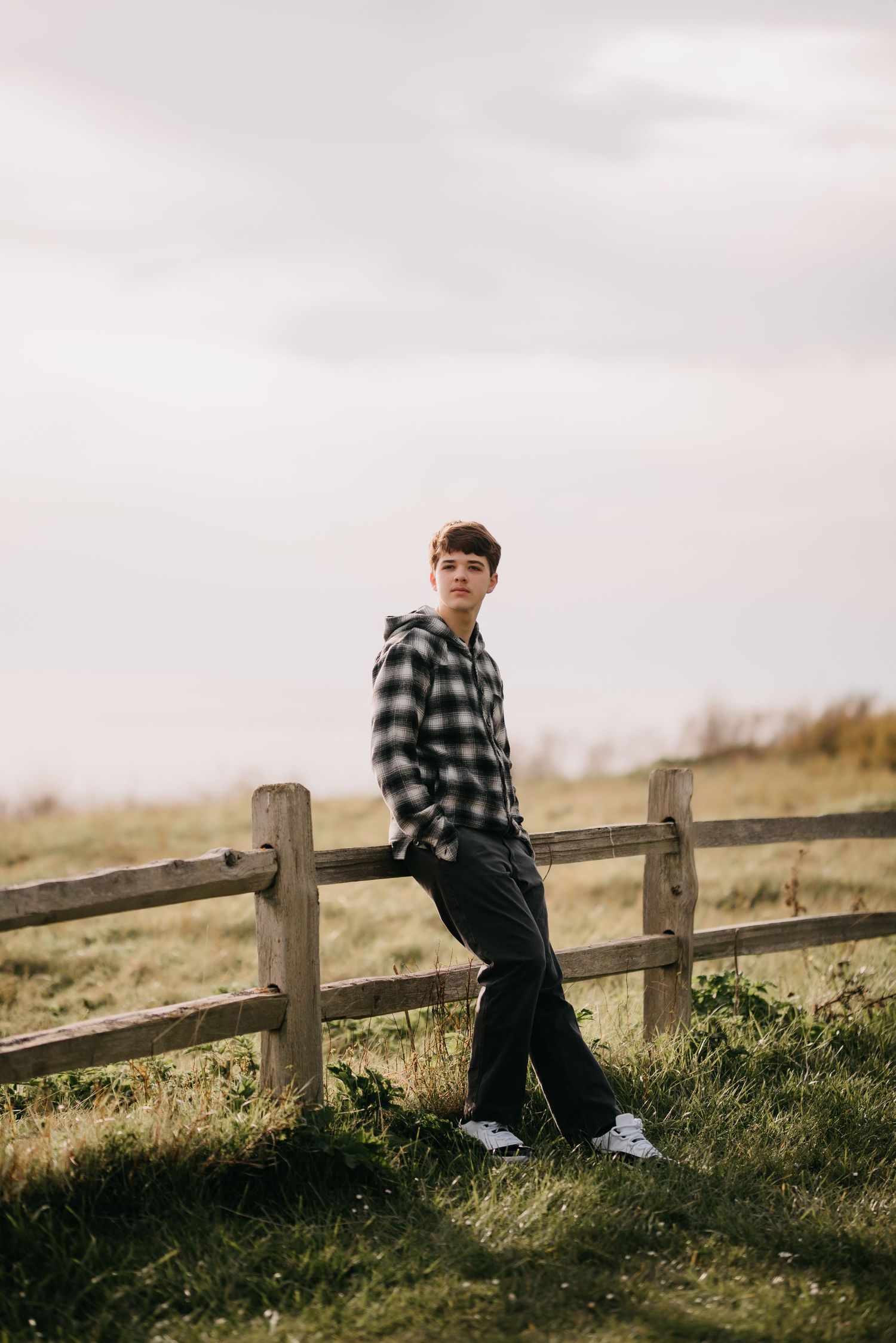 Young man in black and white plaid hoodie leaning against a wooden fence in a green field.