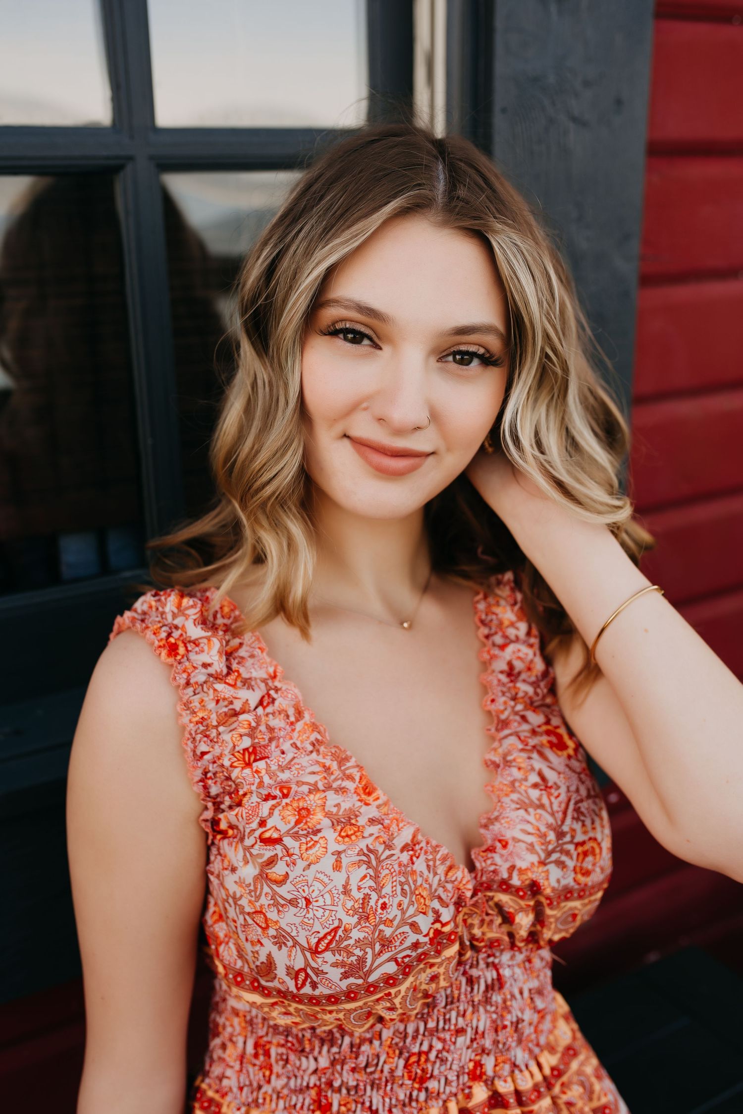 Young woman with wavy blonde hair smiles in floral orange dress for senior portrait outdoors.